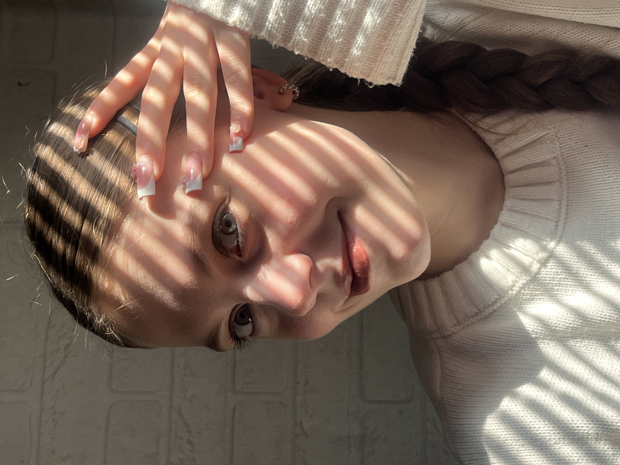 A portrait of a young woman with light skin, brown hair in a braid, wearing a white knitted sweater. She has light-colored eyes and is wearing lipstick. Her left hand is raised near her head, revealing manicured nails with floral accents. Sunlight streams through what seems to be window blinds, casting alternating shadows and light stripes across her face and hand. The background features a white brick or textured wall. The location cannot be determined from the image.