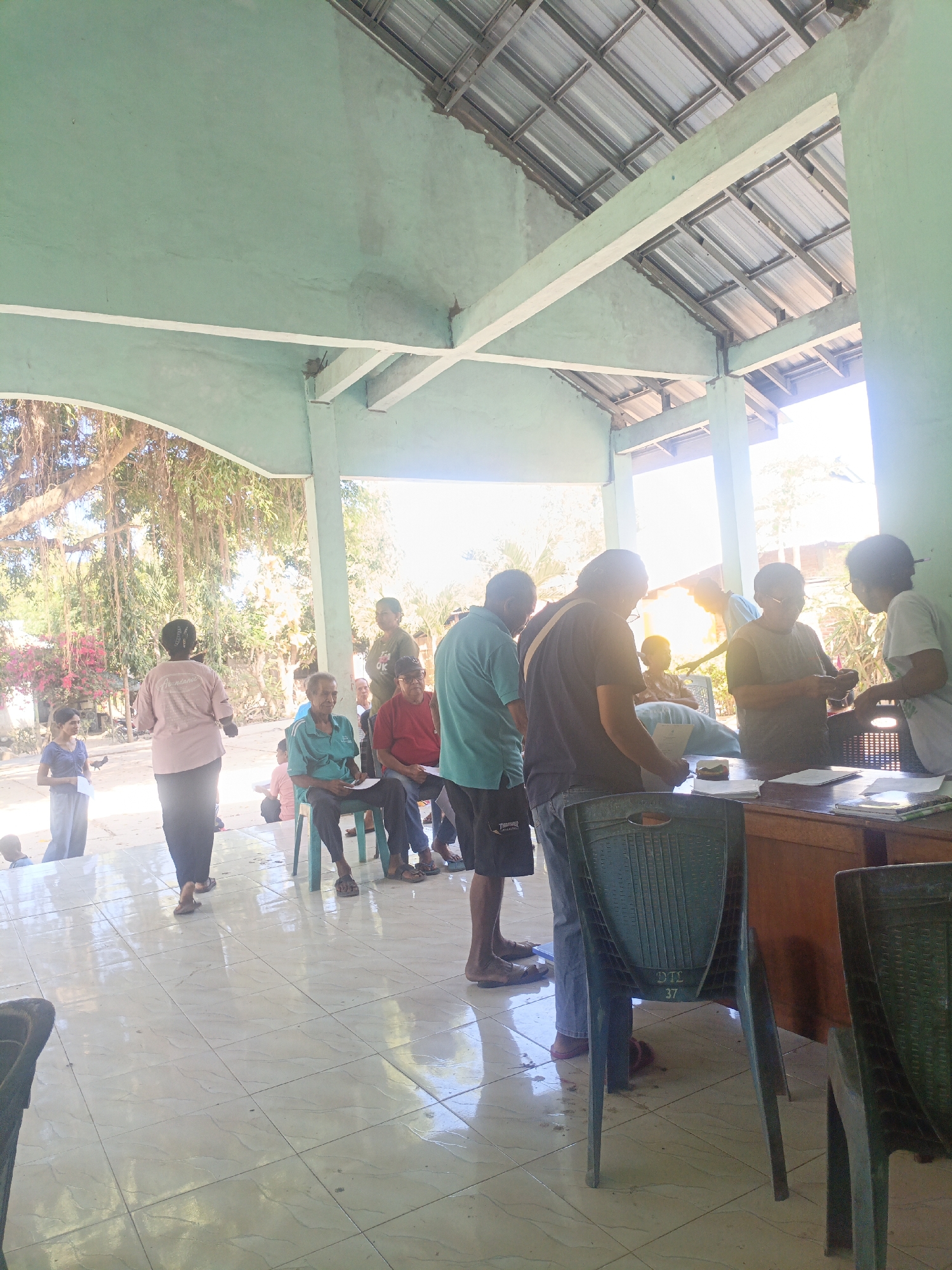 In a covered outdoor area with a light green painted ceiling and metal roof, several individuals are gathered. The ground is tiled with light-colored square tiles that reflect the sunlight. On the right side of the frame, a wooden desk is visible with papers and books on it, suggesting an administrative or registration setting. Several dark green plastic chairs are placed around the desk and in the open area.
In the foreground, a dark green plastic chair is prominently featured, with the markings "DTL 37" visible on its back. Further into the scene, a group of people are standing and sitting, some appearing to be interacting with each other. One individual in the foreground, wearing a blue shirt and dark shorts, is looking towards the desk. Another person in a dark shirt and jeans is also near the desk. Further back, a woman in a pink shirt and dark pants is facing away from the camera, appearing to be addressing someone. Several other individuals are seated on chairs, holding papers.
The background reveals an outdoor environment with trees, including one with prominent hanging roots, and pink flowering bushes. The overall impression is of a public gathering or service point in a rural or semi-rural setting, possibly in the city of Tanah Lein, Indonesia, given the context. The bright sunlight indicates it is daytime.