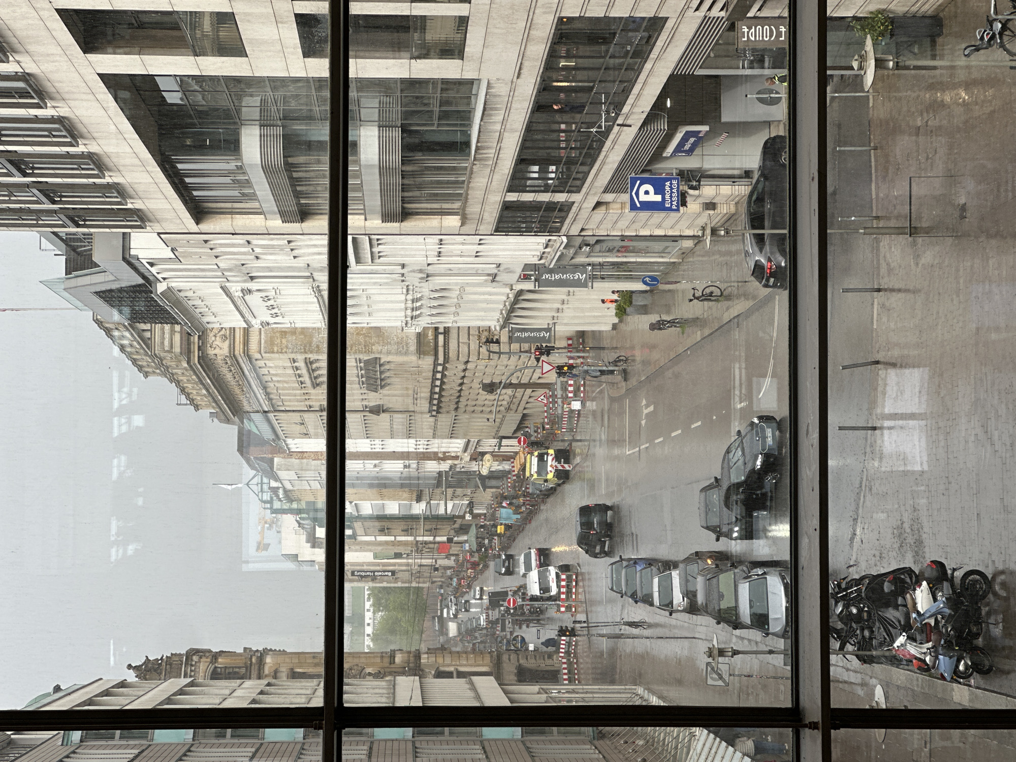 This image presents an overhead view of a bustling city street in Hamburg, Germany, observed through a window. The scene depicts a typical urban environment on an overcast or rainy day, with wet pavement reflecting light, suggesting recent or ongoing precipitation.
The street is lined with a mix of architectural styles, from modern buildings with glass facades and clean lines to older, more ornate structures featuring classical detailing. Numerous vehicles populate the street, including cars, motorcycles, and scooters, some parked along the curb and others navigating the traffic. A white utility van is also visible, and red and white striped construction barriers suggest road work or an event taking place on one side of the street.
Notable details include reflections on the window through which the photo was taken, partially obscuring the view but also adding depth. Street signs, traffic lights, and street furniture are visible throughout.
Several pieces of text are discernible:
- A blue parking sign with a white "P" and the words "EUROPA PASSAGE".
- A banner on a building reads "Barceló Hamburg".
- The name "hessnatur" appears on the facades of at least two buildings.
- A partially visible and reflected sign reads "E COUPE" or "F COUPE".
- Various German road signs are also visible.
A few pedestrians can be seen on the sidewalks, and a person in a yellow vest is visible in a reflection in one of the building's windows. The overall impression is that of active city life continuing despite the gloomy weather.