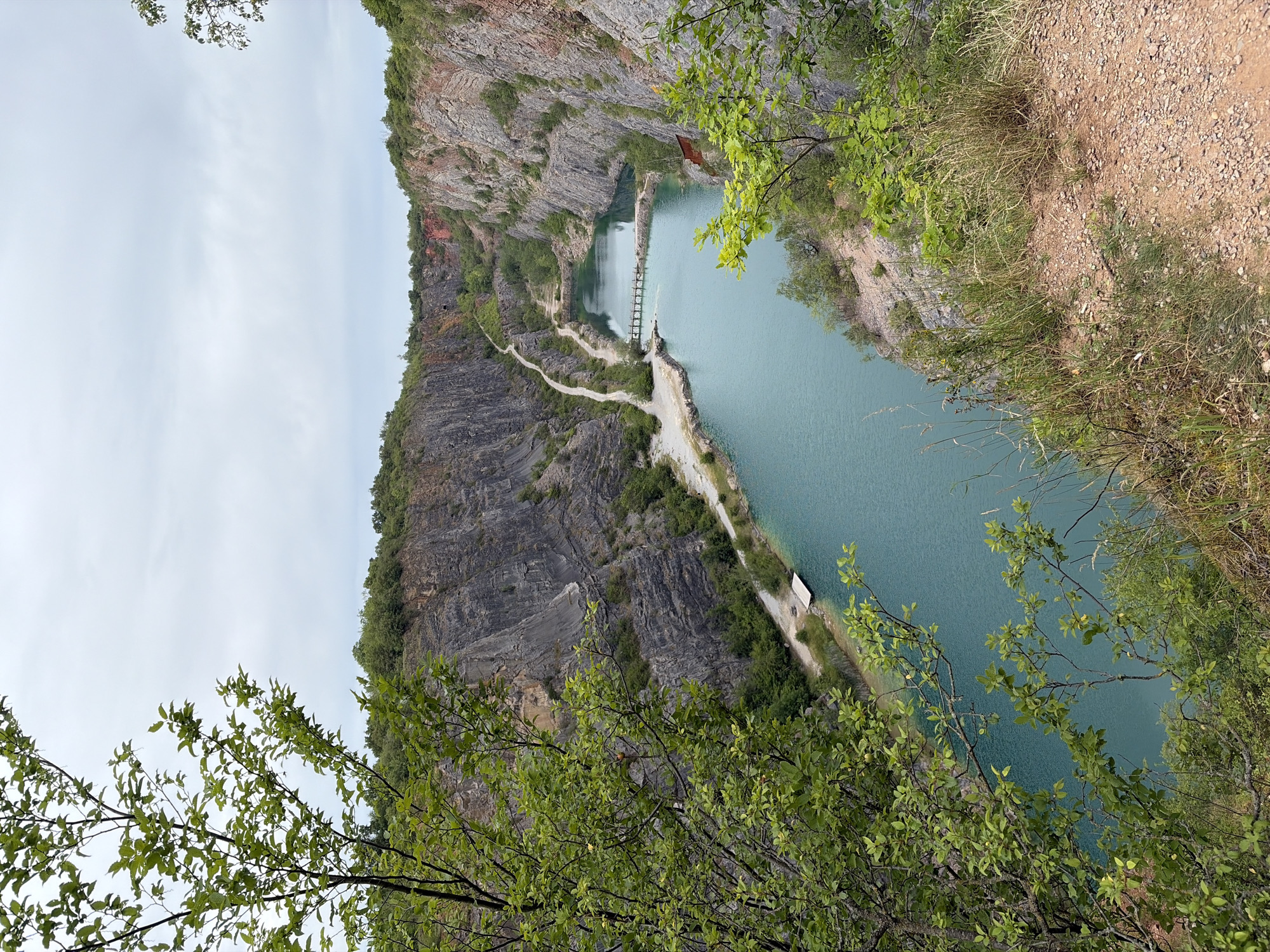 The image shows a quarry in Trněný Újezd, Czechia. The quarry is filled with turquoise water, creating a striking contrast with the surrounding rocky cliffs. A wooden footbridge spans across the water in the middle of the quarry. A dirt path winds along the edge of the quarry, with some vegetation growing on the slopes. The sky is overcast, creating a subdued light over the scene. In the foreground, a leafy tree branch partially obscures the view, adding depth and framing the landscape.