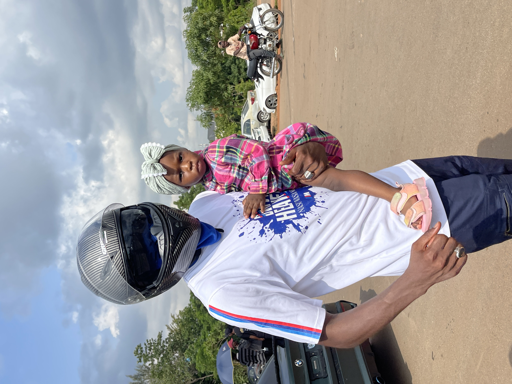 A man wearing a black motorcycle helmet and a white t-shirt with blue and red stripes on the sleeve holds a young child dressed in a pink plaid outfit. The child wears a light green headband. The man's t-shirt has a blue splatter graphic with text that reads "UNLIMITED HEART" and "NASU VSAN". He is also wearing dark blue pants. In the background, several cars are parked, including a white convertible and a dark colored car with a BMW logo on its trunk. A man on a red motorcycle is visible further in the background. Trees and buildings are present in the distance, under a cloudy sky. The scene appears to be an outdoor gathering or event.