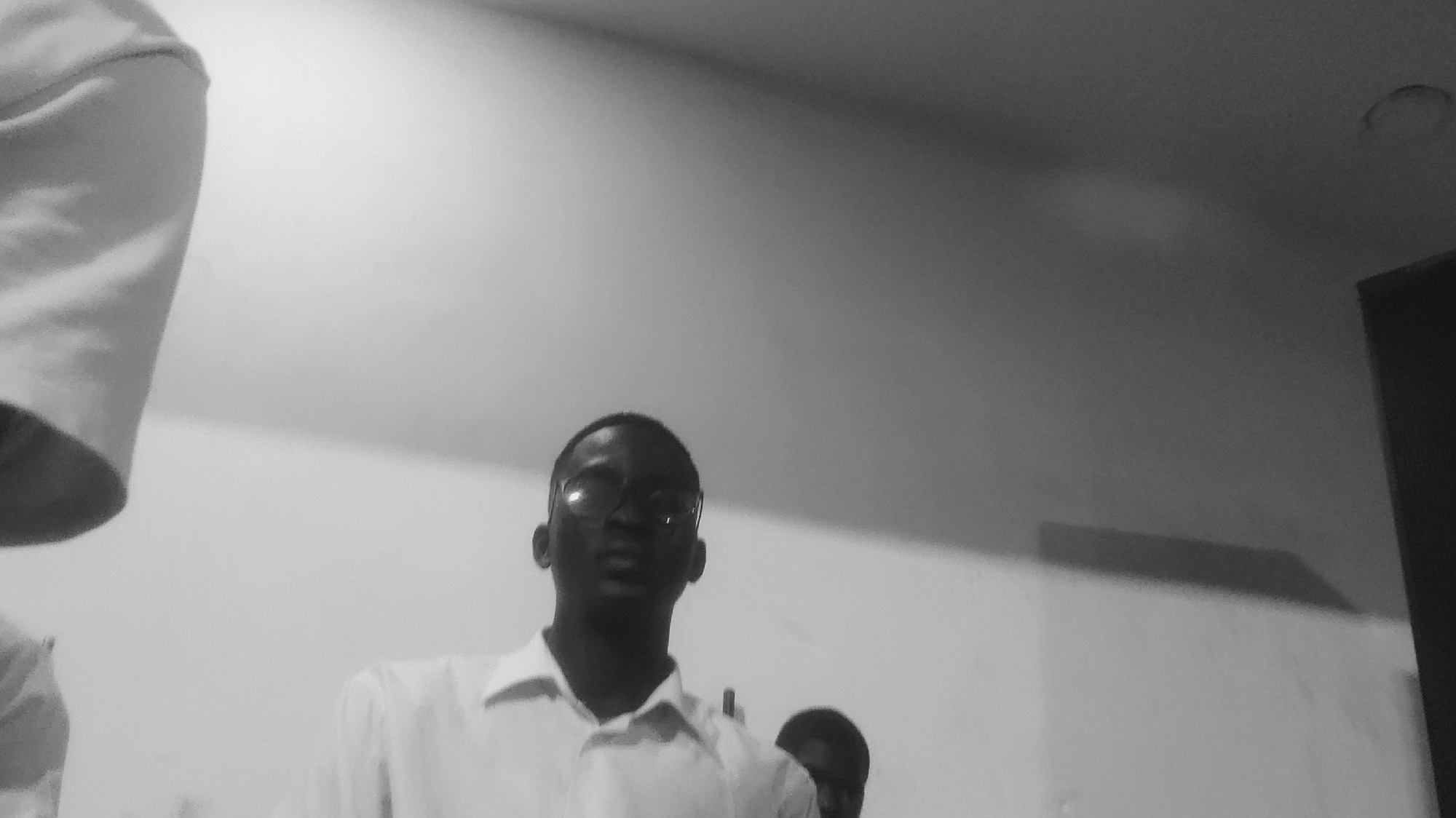 A black and white, low-angle shot shows two young men in what appears to be an indoor setting, possibly a classroom or meeting room, in Jalingo, Nigeria. The man in the foreground, positioned slightly to the left of center, is wearing glasses and a white collared shirt. His head is tilted slightly upwards, and he is looking directly at the camera. To his right and slightly behind him, another young man with a shaved head is visible, also wearing a white shirt. The background consists of a plain, illuminated wall and a ceiling. The lighting suggests it might be daylight, with a light fixture possibly visible on the ceiling. The overall impression is candid, with a focus on the individuals in the frame.