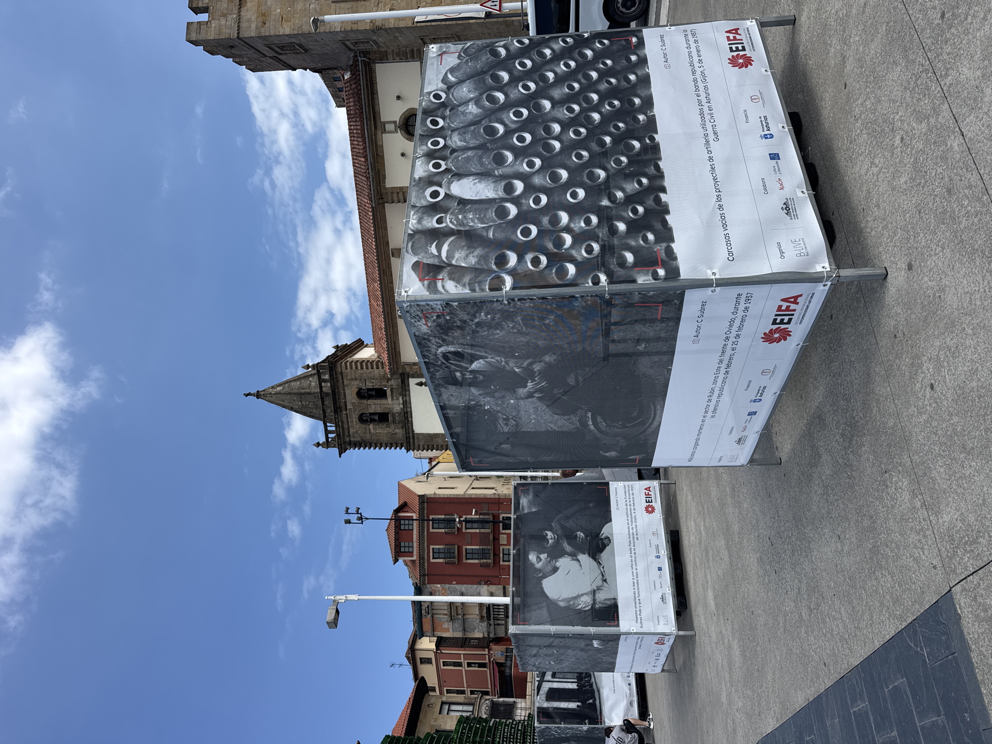The image captures an outdoor exhibition in Gijón, Spain, with historical photographs displayed on large, fabric-covered structures. In the foreground, a textured concrete plaza is visible, with a section of dark grey stone paving on the left. Three large, rectangular display structures are the primary focus. The first, on the left, shows a black and white photograph of a woman and child. The second, in the center, displays a close-up of rows of artillery shell casings. The third, on the right, also features artillery shell casings. White banners with text and logos are attached to the bottom of these structures. Text on the banners includes "EifA" and "ENCUENTROS FOTOGRÁFICOS ASTURIAS." In the background, several multi-story buildings with red-tiled roofs and colorful facades stand under a bright blue sky with scattered clouds. A church tower with a stone steeple is also visible. A white lamp post is positioned to the left of the central display. A dark-colored car is parked behind the display on the right. Two people are partially visible in the image: one person is seated on the ground in front of the leftmost display, and another person's legs are visible beneath the display on the right. The overall scene suggests a cultural event or exhibition taking place in a public urban space.
