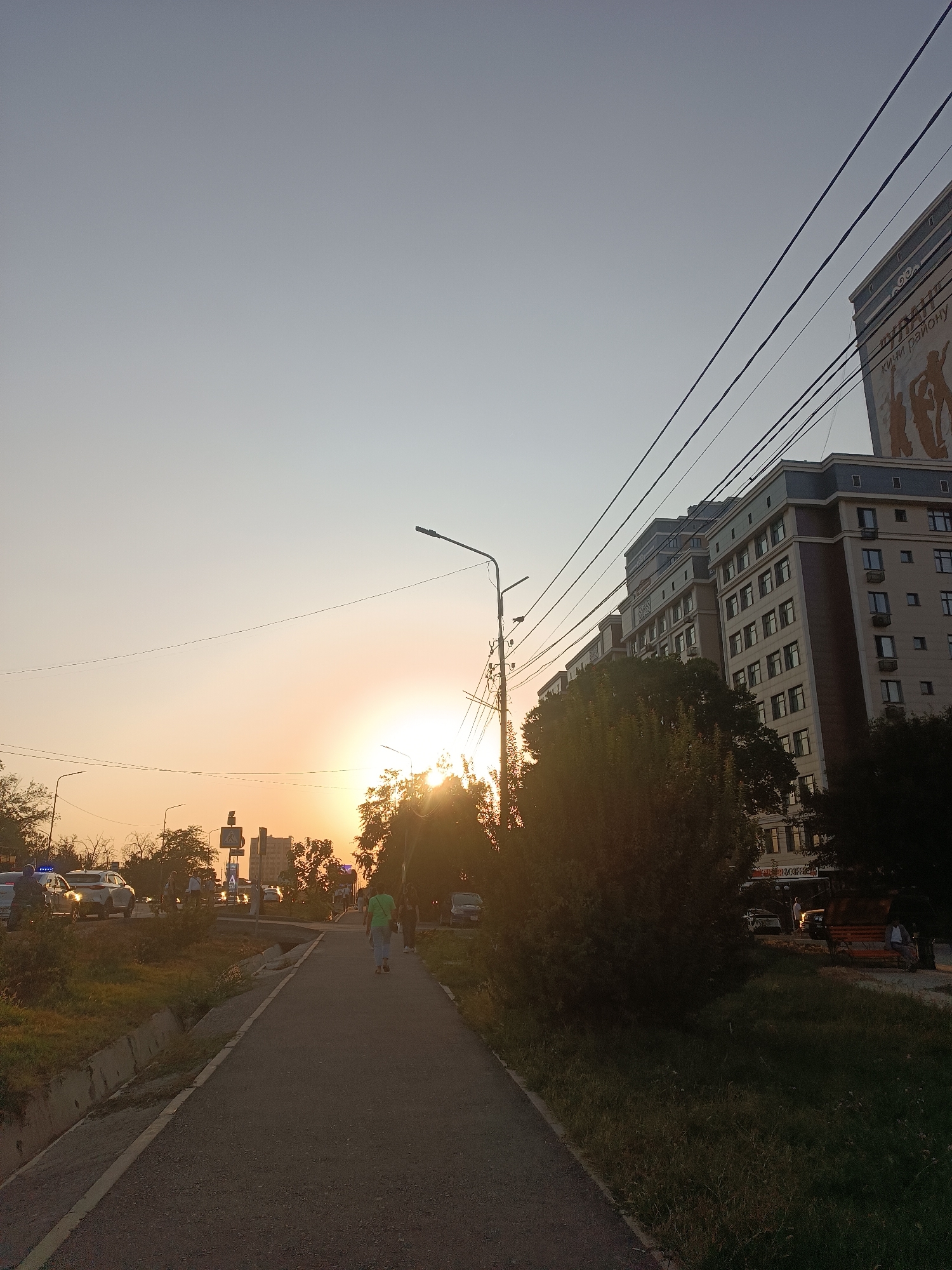 The image depicts a street scene in Bishkek, Kyrgyzstan, during sunset. A pedestrian walkway runs along the right side of the road, with a grassy area and a drainage ditch to its left. A building with multiple floors and windows is visible on the right, with power lines stretching across the sky in front of it. In the distance, traffic can be seen on the road, including cars and a police car with its lights on. Trees line the street, and the setting sun casts a warm glow over the scene. A few pedestrians are walking on the walkway, and one person is sitting on a bench in the foreground. The overall impression is one of an urban environment at dusk, with the natural beauty of the sunset adding a touch of serenity.