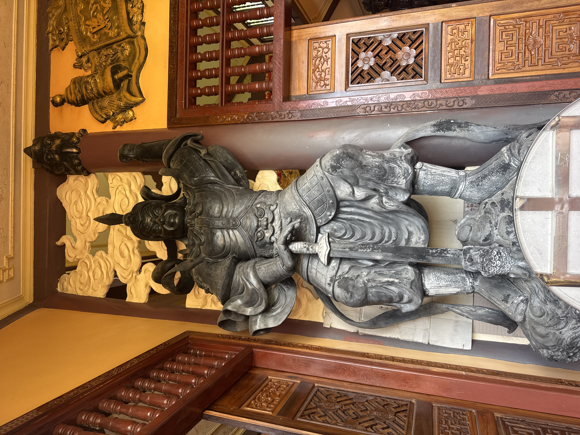 This image captures a large, imposing statue of a warrior, likely a Buddhist deity or protector, situated in a temple or cultural site in Da Lat, Vietnam. The statue is made of dark, weathered material, possibly bronze or stone, and depicts the figure in full armor, with a stern expression, a helmet, and holding a sword. The warrior is in a dynamic pose, with one arm raised and the other resting on the sword hilt.
The statue is positioned against a backdrop of ornate architectural elements. To the left and right are intricately carved wooden doors and latticework, typical of Vietnamese traditional architecture. The colors of the surrounding walls are a vibrant yellow, contrasting with the dark statue and the reddish-brown wood. Behind the warrior, there's a carved panel featuring stylized clouds. Above and to the right, a golden relief adorns the wall, depicting what appears to be a scroll and some ceremonial objects. The overall impression is one of grandeur and reverence.
The scene is illuminated by natural light, casting subtle shadows that emphasize the texture and details of the statue and the surrounding decor. The floor area in front of the statue is partially visible, appearing to be a tiled surface with some metallic elements. The statue is placed on a base that is sculpted to look like a rocky outcrop or a mythical beast, further adding to its imposing presence. The overall atmosphere conveyed by the image is one of cultural richness and historical significance.