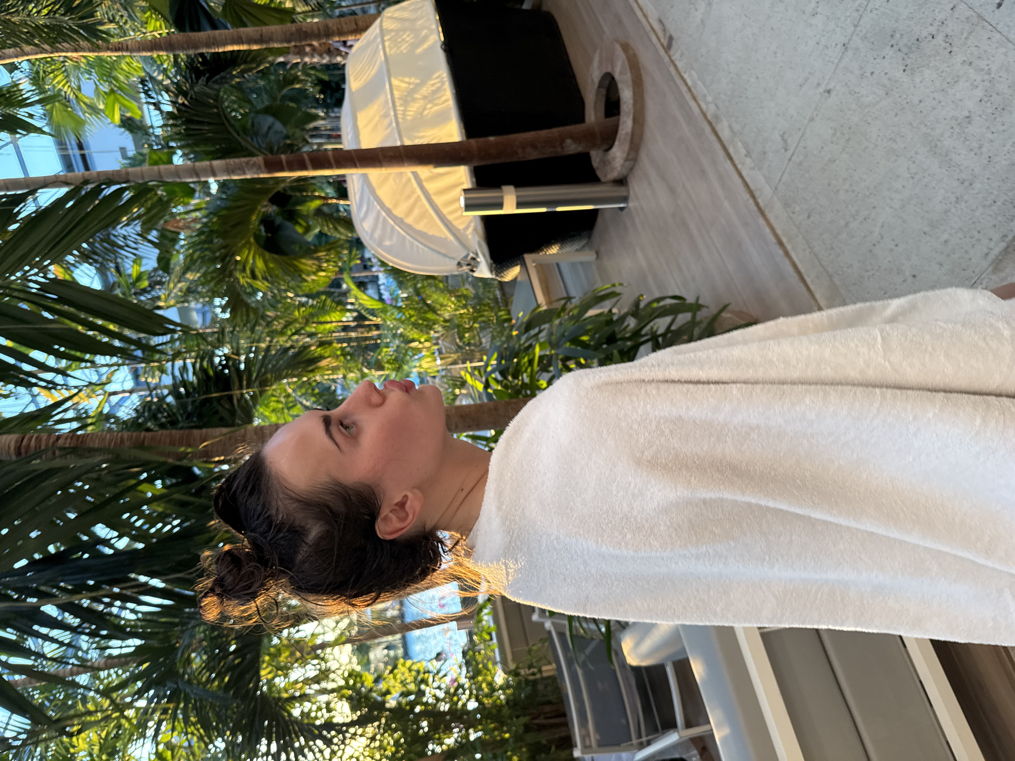 The image depicts a woman wrapped in a white towel, likely a spa or hotel guest. She is looking upwards in a setting with lush tropical foliage, possibly palm trees, visible in the background. There is a sunbed or lounge chair nearby. The scene appears to be outdoors, potentially in a resort or spa environment. The exact location is not possible to determine with certainty, but the presence of tropical vegetation suggests a warm climate.
