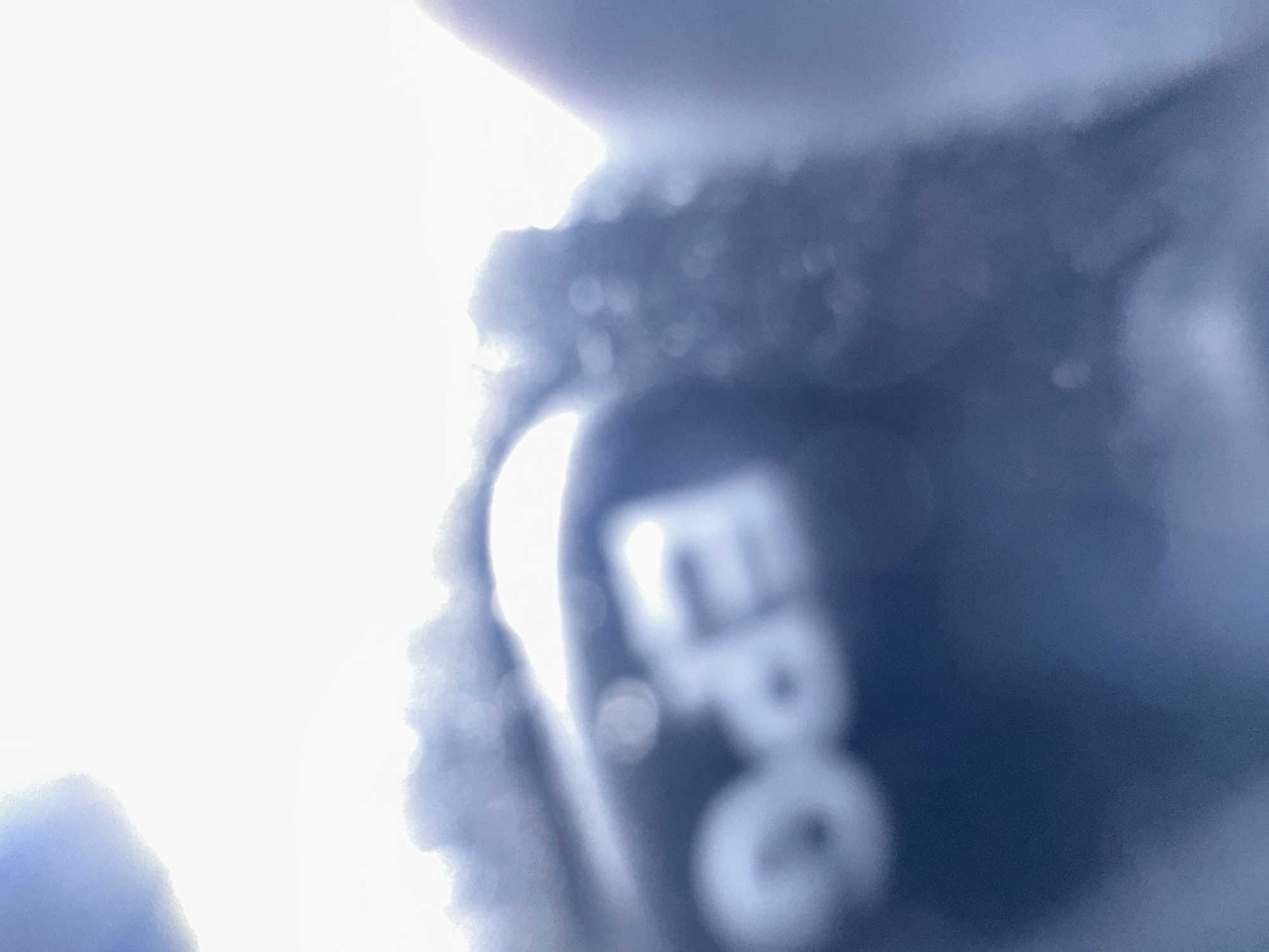 The image is a close-up, out-of-focus shot of a dark object with the letters "EPG" visible in a lighter color. The object appears to be made of a textured material, possibly fabric or rubber, and is illuminated by a bright light source from the left, creating a strong glare and obscuring finer details. The overall tone of the image is dark blue and white. The "EPG" text is partially obscured by glare and the blurriness of the image, but the letters are discernible as E, P, and G. There are also some circular highlights, likely reflections or bokeh, scattered throughout the image. The setting is unclear due to the extreme close-up and lack of context, but the bright light suggests it could be outdoors or near a strong artificial light source. No specific activity or event can be identified. The location context of Belgrade, Serbia does not provide any specific clues for this abstract image.