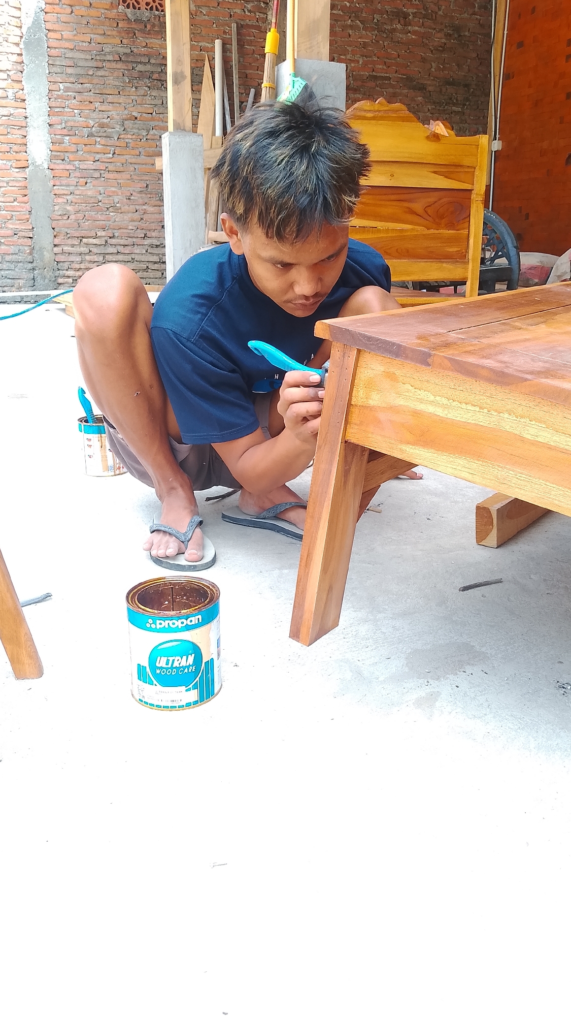 In Karanglangu, Indonesia, a young male crouches on a concrete floor, diligently applying wood stain to a wooden table with a bright blue brush. The table, made of light-colored wood, exhibits a polished sheen. To the left of the male, a can of "PROPAN ULTRAN WOOD CARE" is visible, along with another open can and brush. In the background, a brick wall and unfinished wooden structures suggest an outdoor workshop or construction site. The scene is lit by natural light, casting soft shadows on the ground. The overall impression is one of focused work in a rustic setting.