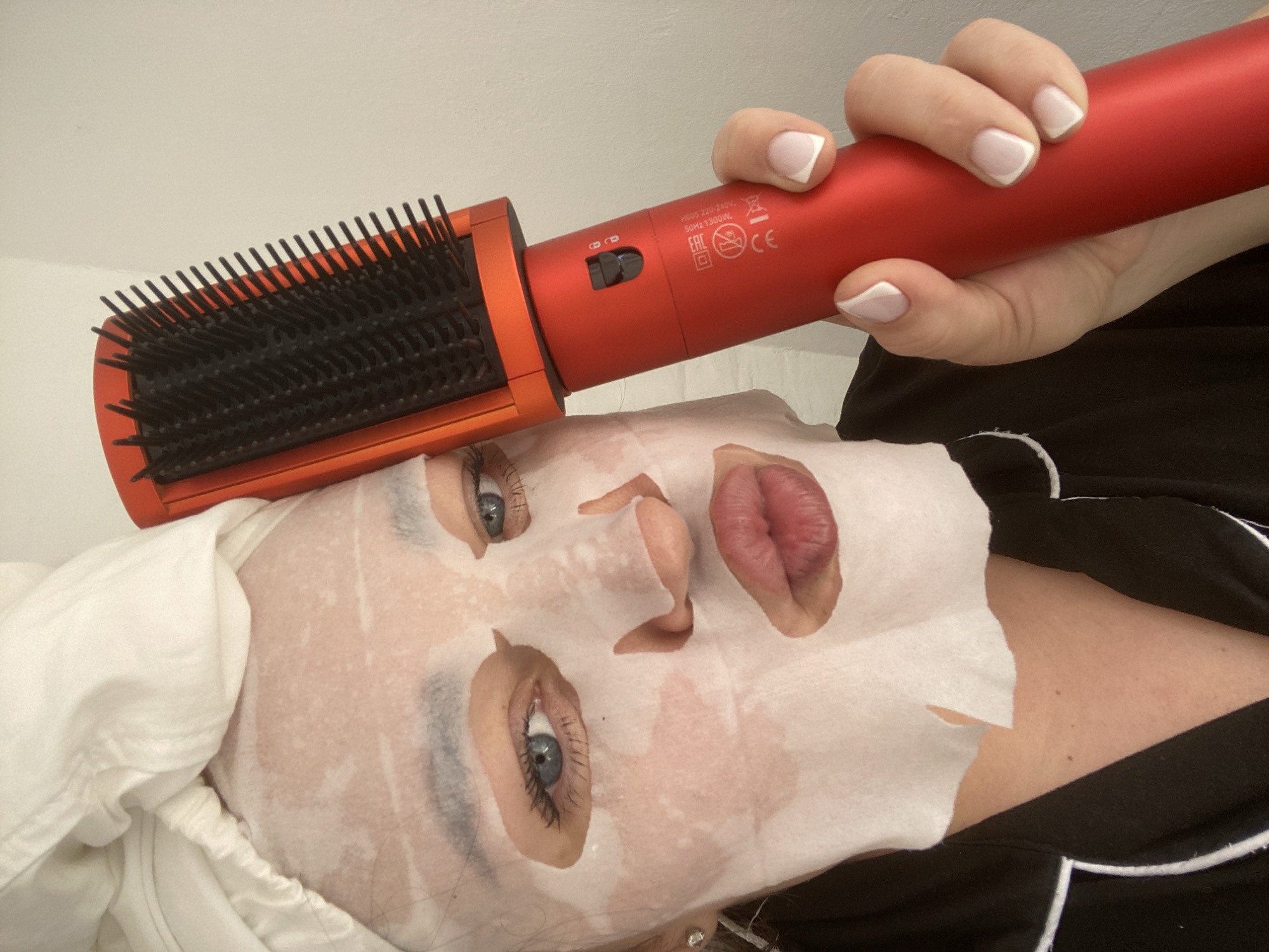 A woman lies on her back wearing a white sheet mask with cutouts for the eyes, nose, and lips. The hair is wrapped in a white towel. Above her, a hand holds a red hair styling brush. The brush has black bristles and an orange head. The hand has a French manicure. The woman is wearing black and white pajamas. The background is a plain white wall. The image does not contain information indicating it was taken in Sochi, Russia.