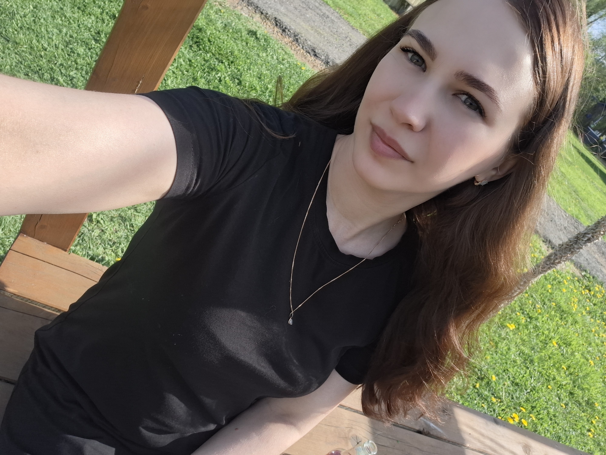 A woman is sitting on a wooden structure, likely a swing or bench, outdoors. She is wearing a black t-shirt and a delicate gold necklace with a pendant. Her hair is long, brown, and wavy, styled off to one side. Her skin is fair, and she has light eye makeup. She is looking directly at the camera with a slight smile.
The background consists of green grass and a pathway or road surface. Yellow dandelions are visible in the grass. Parts of a wooden structure are visible, which could be a support beam or part of the seat itself. The angle of the shot suggests the woman is holding the camera at arm's length, taking a selfie. The presence of grass and trees suggest this is a natural environment.
There is no information in the photo to confirm the specific location is Malyi Bukor, Russia.