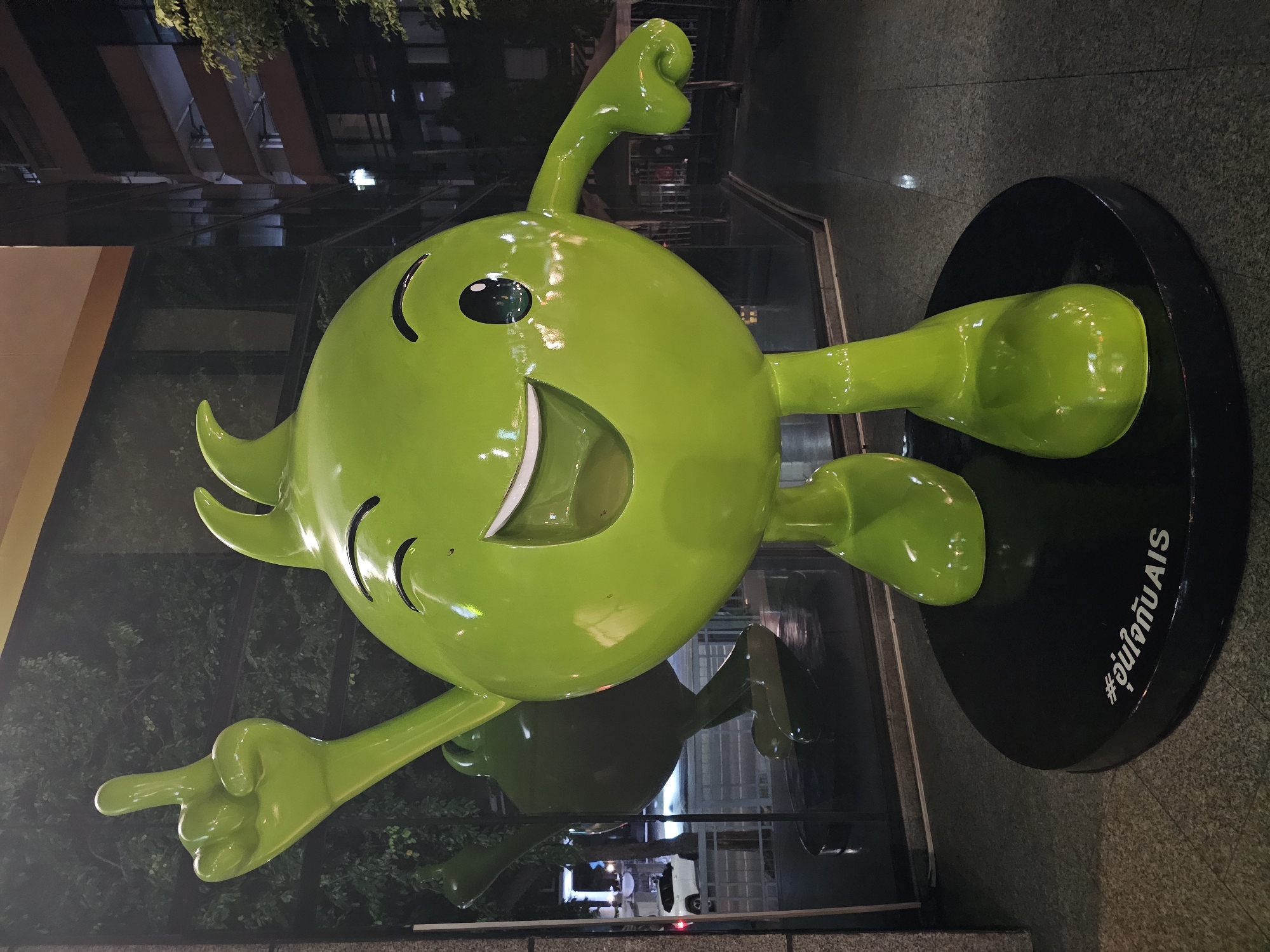 A bright green, cartoonish character sculpture stands on a black circular base in an outdoor urban setting at night. The sculpture is round with a large head and body, two arms, and two legs. It has a wide, smiling mouth, one closed eye, and one open eye with a reflective pupil. The character is pointing its left index finger upwards. The base of the sculpture has white text that appears to be in Thai script. In the background, a glass building with reflective windows and balconies is visible, along with some trees and streetlights. The ground in front of the sculpture is paved with textured tiles. The scene suggests a promotional display or public art installation in Bangkok, Thailand, likely outside a commercial building.