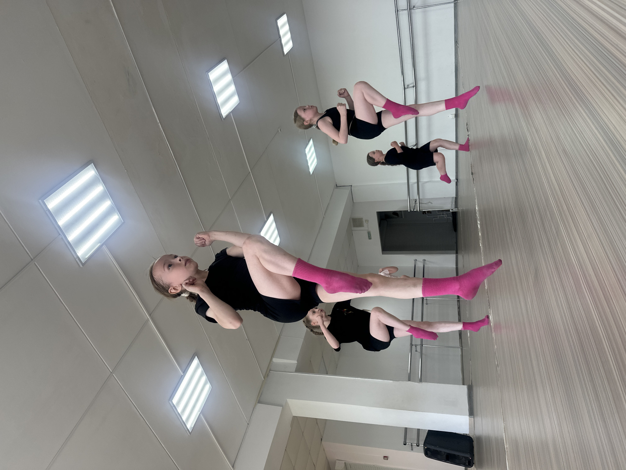 Two young female dancers in black leotards and pink socks are performing exercises in a dance studio. The studio has mirrors, a ballet barre, and fluorescent lighting. The location may be Votkinsk, Russia, but the image does not have enough evidence to confirm this detail.