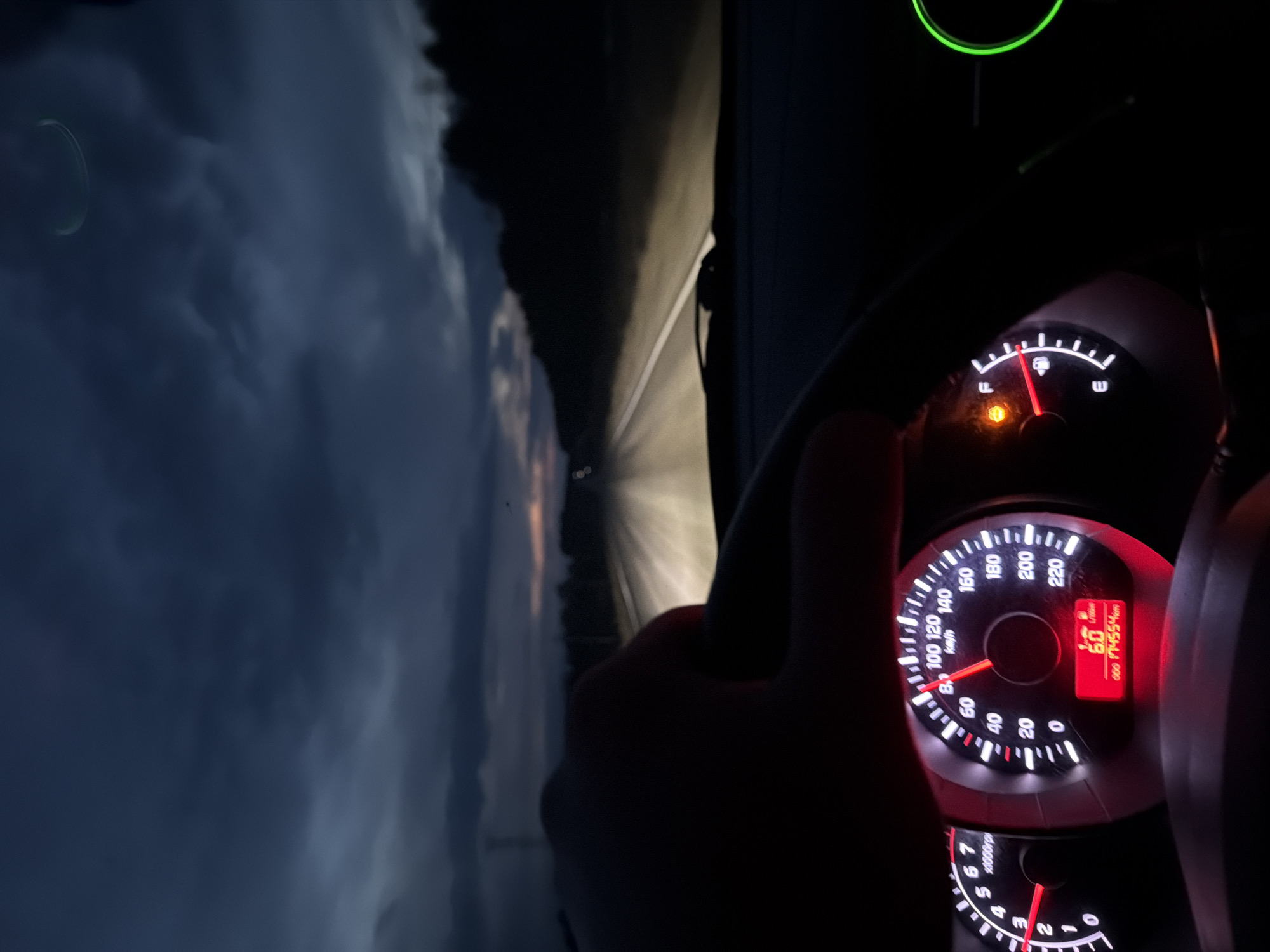 The image shows a car's dashboard at night. The speedometer reads 60 km/h, and the odometer shows 174554 km. The fuel gauge is near empty. The driver's hand is on the steering wheel. The view through the windshield shows a dark road ahead with faint headlights in the distance and dark clouds above. The location is not definitively identifiable as Maksimovo, Russia, based solely on this image.