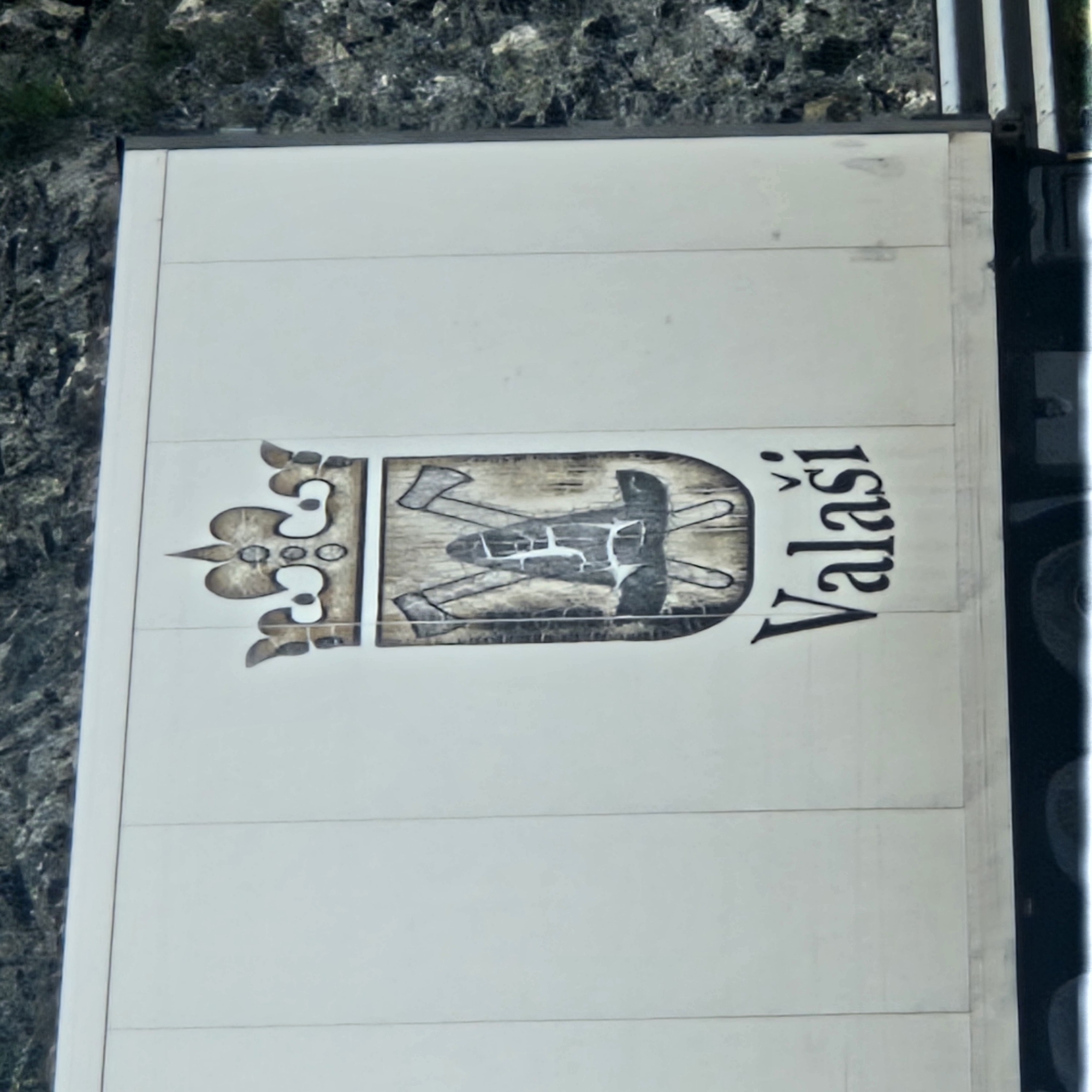 The image displays a large, white rectangular panel, likely the side of a truck trailer, set against a rugged, rocky background with sparse green vegetation. The panel features a distinctive emblem and text.
The emblem, rendered in muted shades of brown, beige, and black, is composed of two main parts. On the left is an ornate, crown-like design with decorative curves and circular elements. To its right, a shield-shaped element contains an illustration of two crossed axes and a third tool resembling a pickaxe or mattock, all depicted with a rustic, textured appearance, including a prominent cross-hatch pattern on the central tool.
Immediately to the right of the emblem, the word "Valaši" is written in bold, black, stylized letters, with a caron (ˇ) above the first 'a'. The sign itself appears to be made of multiple horizontal sections. On the right side of the panel, dark, vertical structures are visible, consistent with the framework or other components of a vehicle.
The scene is illuminated by bright daylight, suggesting clear weather. There is no human activity or specific event taking place, solely the static view of the sign. The rocky terrain in the background indicates an outdoor setting, potentially a mountainous or hilly area, aligning with the context of Le Perthus, France.