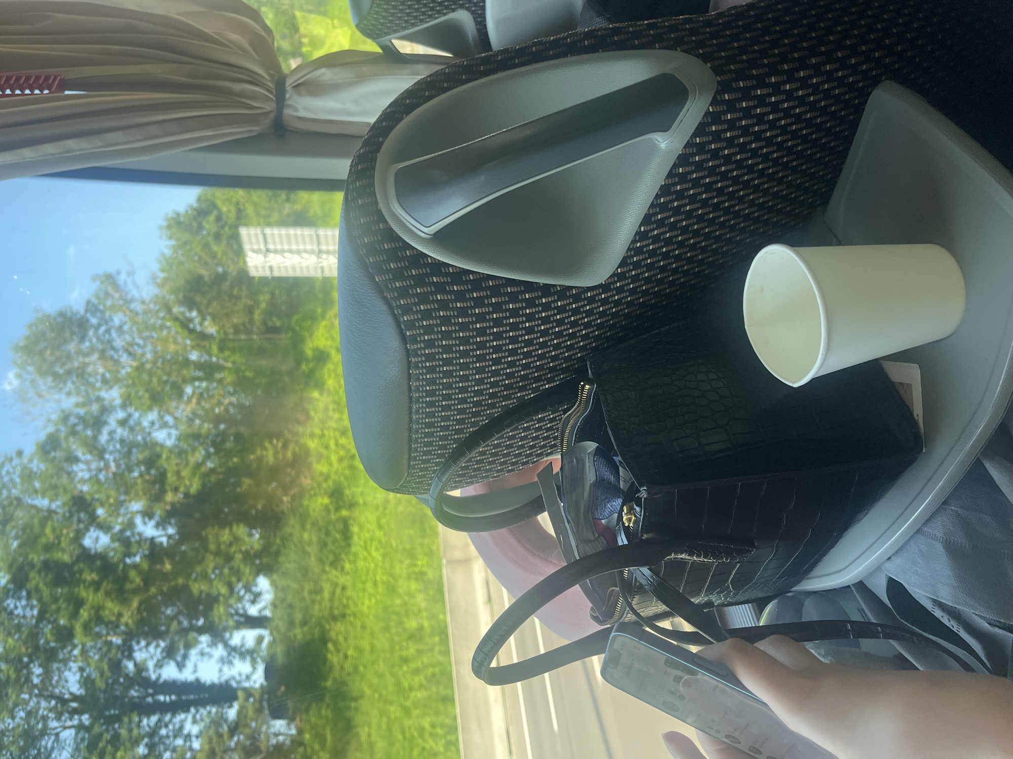 A person's hand, holding a smartphone, is visible on the left side of the frame. In the foreground, a black crocodile-textured handbag with top handles rests on a tray table. Inside the handbag, some items are visible. Next to the handbag on the tray is a white paper cup. The tray table is attached to a seat with patterned fabric and a grey armrest. In the background, a window looks out onto a green, tree-lined landscape under a bright blue sky. The scene appears to be inside a vehicle, possibly a bus or train, traveling through a rural or suburban area. There is no indication of the city of Herstal, Belgium.