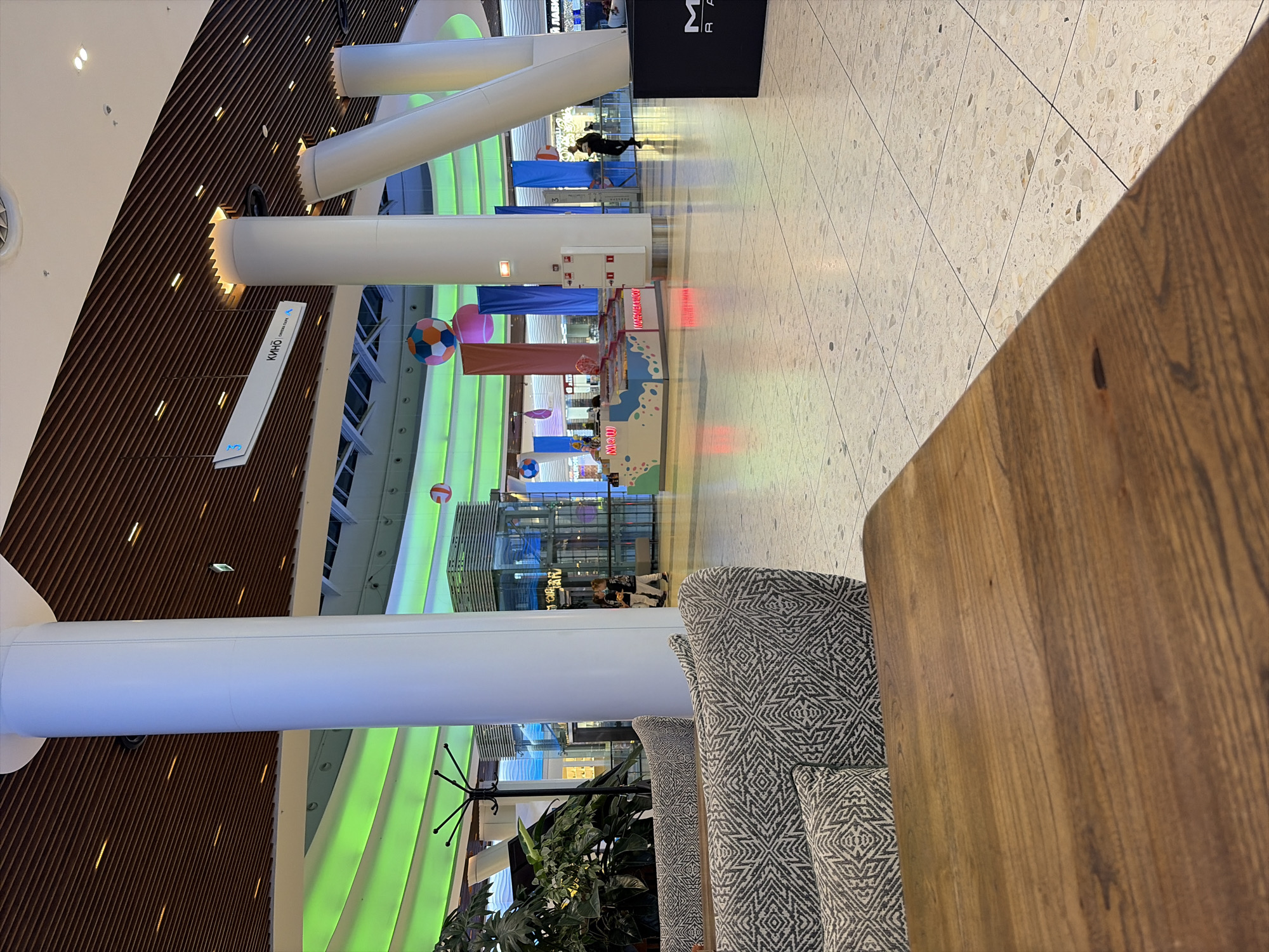 This image depicts an interior scene, likely a shopping mall, in Moscow, Russia. In the foreground, a portion of a wooden table is visible, suggesting a seating area. Behind it, a sofa with a grey and white patterned upholstery is partially shown. The midground reveals large white cylindrical pillars supporting a high ceiling with a brown, slatted wooden finish and accent lighting. Green lighting illuminates curved structures in the background. There are colorful hanging decorations, including spheres and banners, and a glass elevator structure. Several people are present in the midground and background, walking or standing. A kiosk-like structure with a colorful facade is also visible. The floor is tiled with a light-colored, speckled pattern. The overall impression is a modern, brightly lit indoor public space.