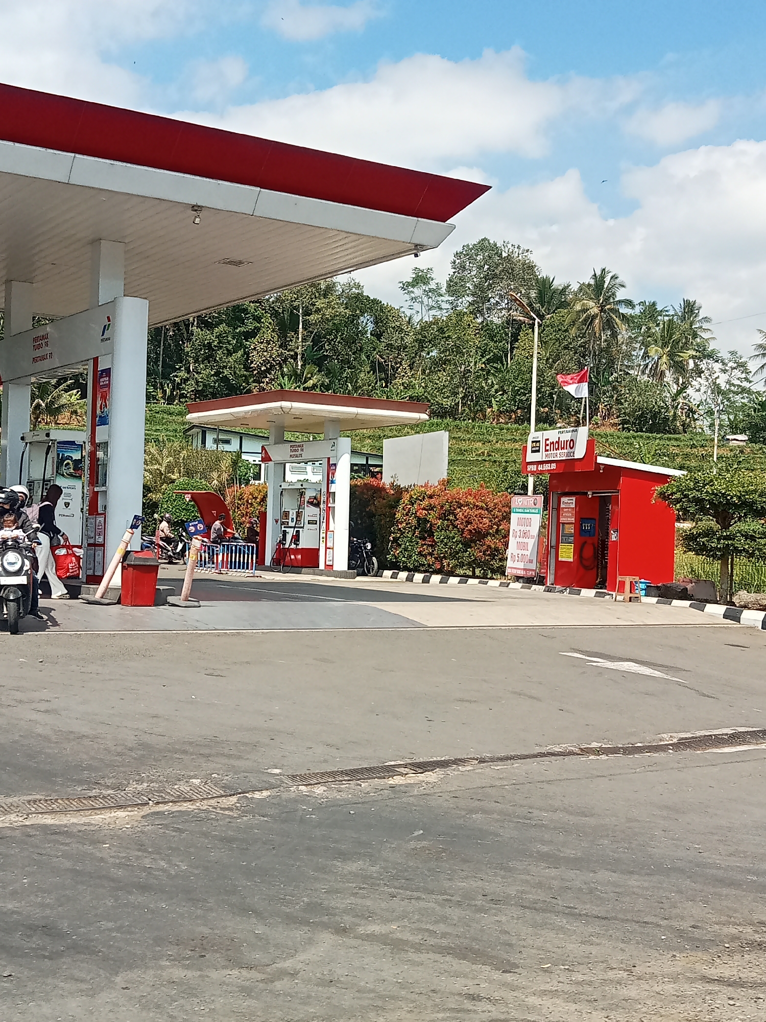 This is a daytime, eye-level shot of a gas station in Selokromo, Indonesia. The station has a red and white canopy over several fuel pumps. To the right, there is a small red building with a sign that reads "Enduro Motor Service". An Indonesian flag is displayed on a pole in front of this building. Several motorcycles are parked or being serviced at the gas station. Two people are visible on a motorcycle in the foreground, and others are seen near the pumps and the service building. The gas station is situated on a paved road with a drain running across it. Behind the gas station, there are lush green trees and vegetation, with a hillside in the background. The sky is blue with some white clouds.