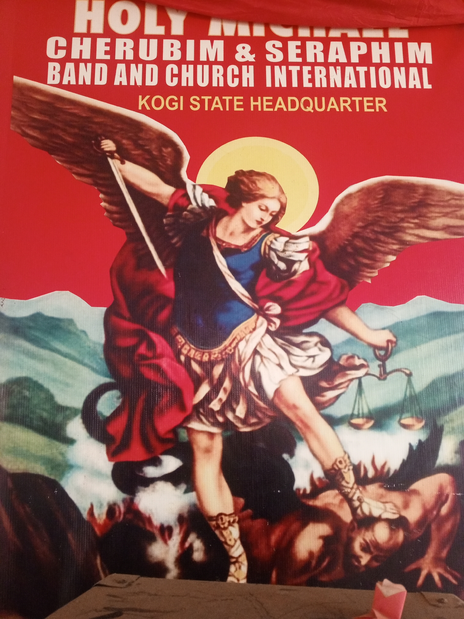 The image depicts a poster with the text "HOLY MICHAEL CHERUBIM & SERAPHIM BAND AND CHURCH INTERNATIONAL KOGI STATE HEADQUARTER" displayed prominently at the top. Below the text is an artistic representation of the Archangel Michael standing over a fallen figure, presumed to be the devil, with his foot on the figure's head. Michael is winged, holds a sword in one hand, and a scale in the other. A halo is positioned behind his head. The background includes a landscape with mountains and foliage. The setting is likely Kogi State Headquarters, Nigeria.