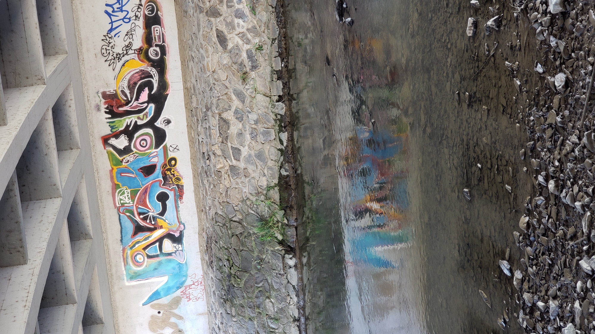 Under a concrete bridge, a section of the concrete wall is covered with vibrant graffiti art. The graffiti features abstract shapes and designs in various colors including blue, red, yellow, pink, black, and white. Below the wall is a stone embankment, and further down, a calm body of water reflects the colors of the graffiti. The water's surface is slightly rippled, and the riverbed is visible, covered with numerous stones. The scene appears to be an outdoor urban environment, possibly a river running through the city of Prešov, Slovakia, given the context of the graffiti art. There are no people visible in the image.