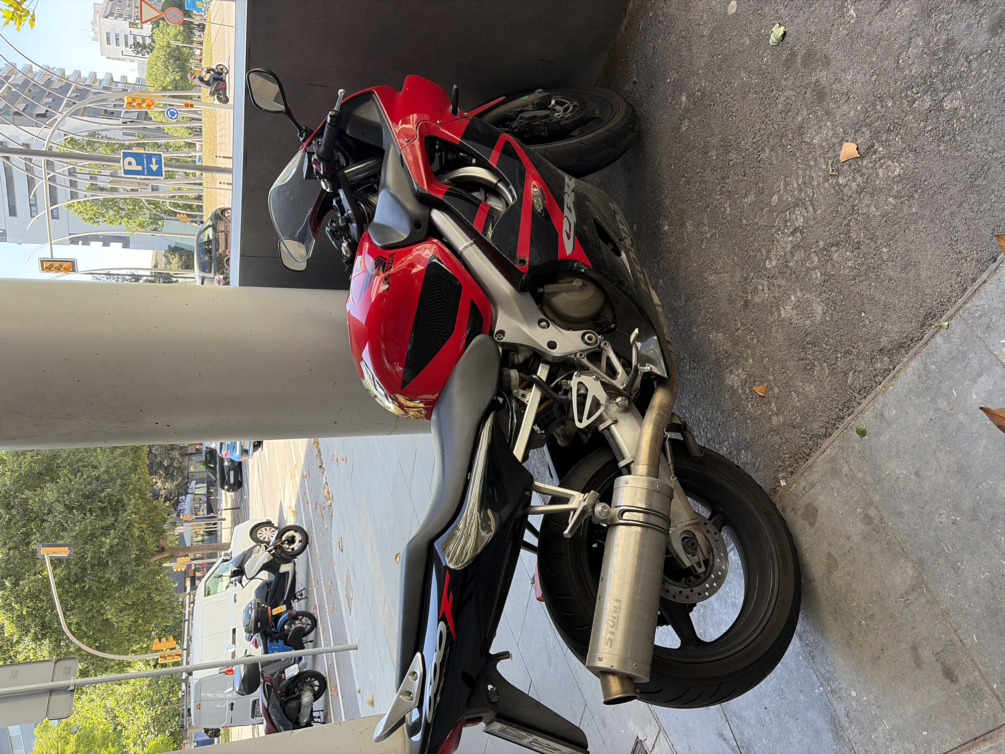 The image shows a red and black Honda CBR 600 motorcycle parked in a covered outdoor area, likely a sidewalk or under a building overhang. The motorcycle is positioned on a grey paved surface. The rear of the motorcycle is closer to the viewer, and the front is pointed towards the back of the frame. A silver exhaust pipe is visible near the front wheel.
Through an opening behind the motorcycle, a street scene can be seen. There are several vehicles parked on the street, including white vans and a blue car. In the distance, some street lights and other motorcycles parked along the sidewalk are visible. There are some trees visible. The architecture in the background includes a modern building with white curved supports. Based on the architectural details and the street signage visible in the background, the location is l'Hospitalet de Llobregat, Spain. No people are clearly visible in the image.