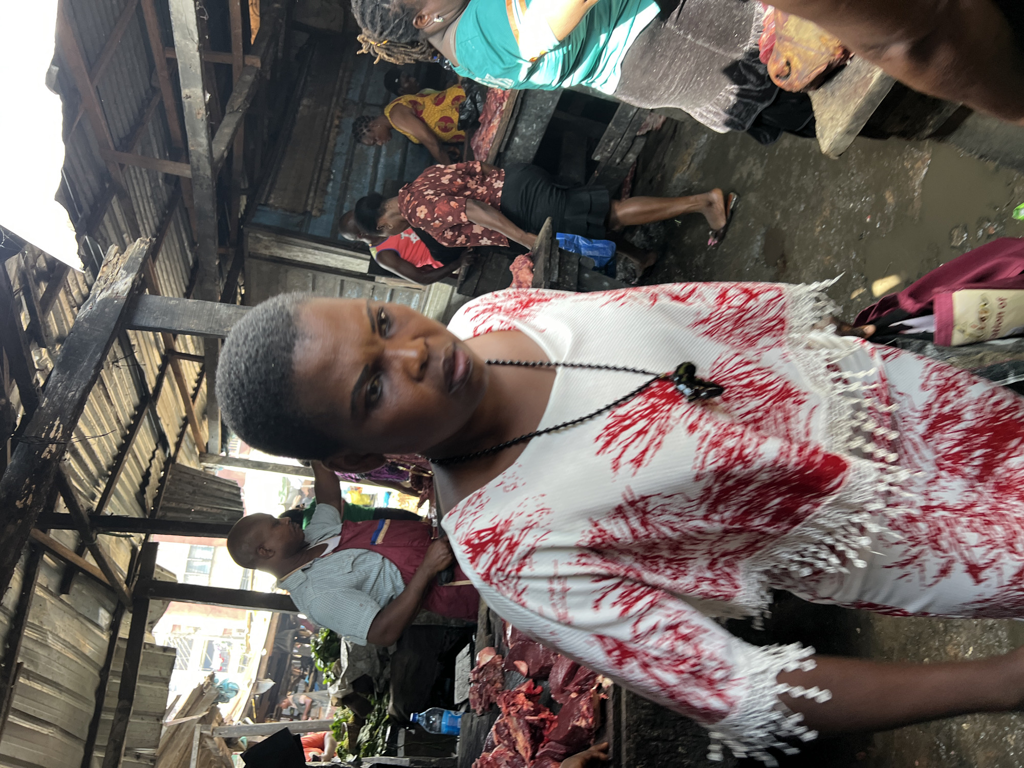 The image depicts a bustling outdoor market scene, likely in Nkpor, Nigeria, characterized by its raw and authentic atmosphere. A woman in the foreground, wearing a white dress with red abstract prints and a beaded necklace, appears to be the main subject. She is captured in motion, her gaze directed downwards with a focused expression. Behind her, a man in a checkered shirt and a red apron is visible, suggesting he might be a vendor. Scattered around the market are stalls with raw meat, fresh produce, and various other goods, indicative of a typical Nigerian marketplace. The environment appears somewhat unkempt, with visible debris and water on the ground, and the structures are makeshift, made of corrugated iron and wood, suggesting an informal economic setting. Other market-goers are seen in the background, engaged in their respective activities, contributing to the lively and dynamic ambiance of the scene. The overall image captures a snapshot of daily life and commerce in a Nigerian urban setting.