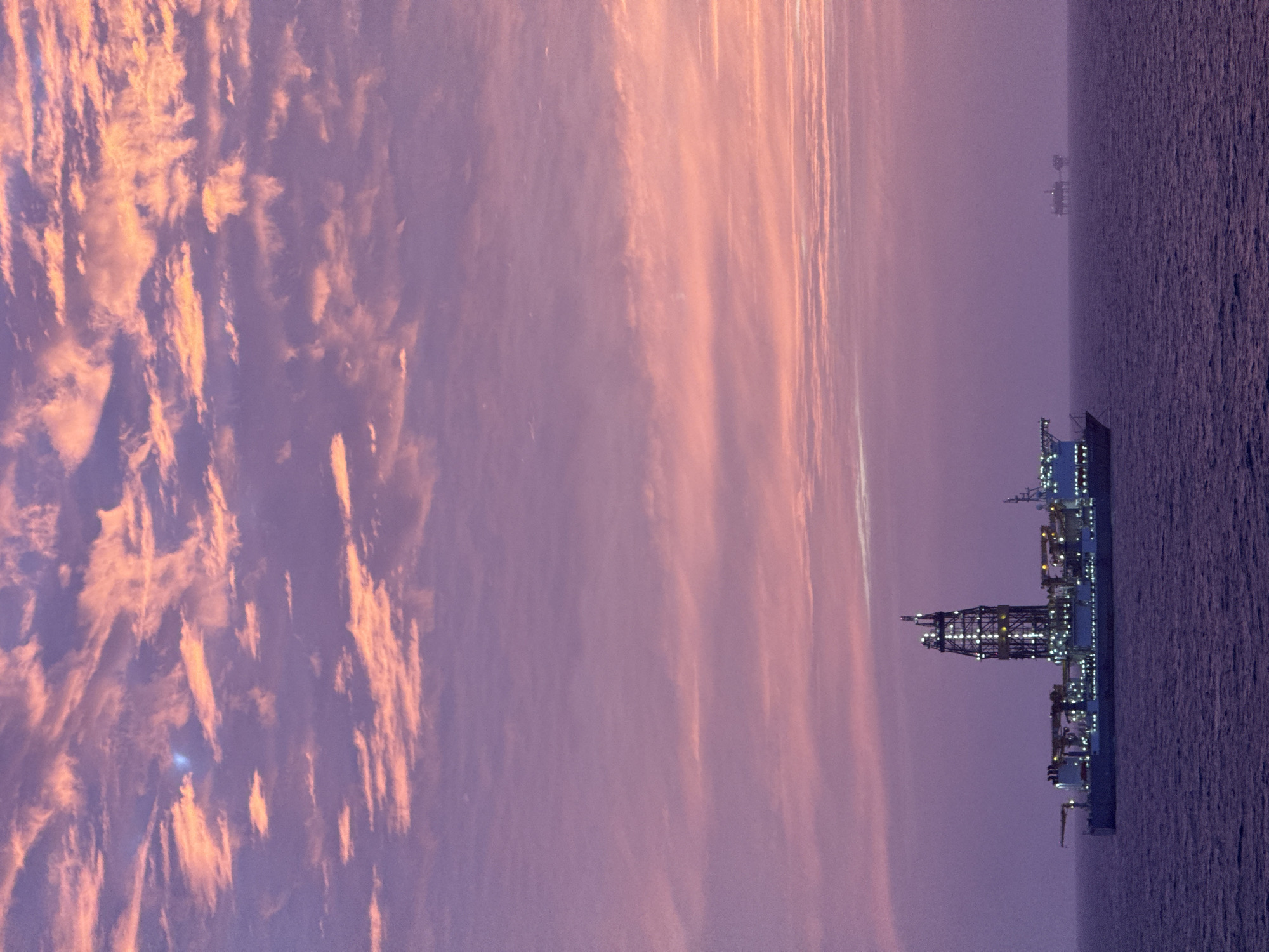 The image shows a large oil platform at sea against a backdrop of a pastel pink and purple sky. Another smaller platform can be seen in the distance. The sky is streaked with clouds. The location is unknown.