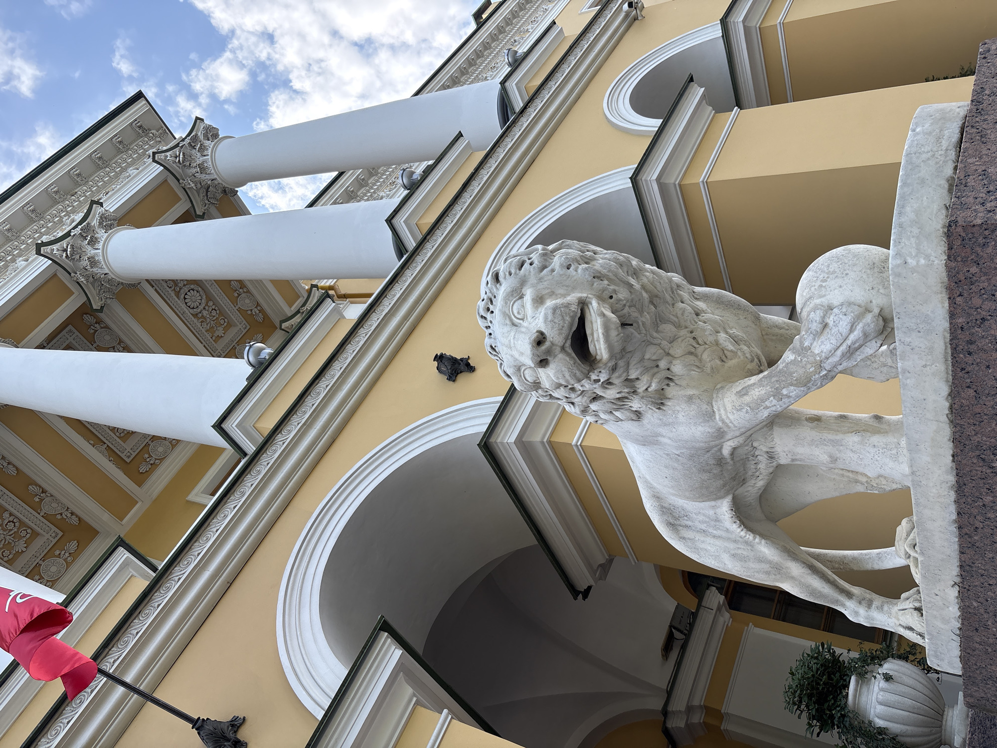 The image shows a white marble lion statue with its mouth open, standing in front of a yellow building with white columns. A red flag is flying on a pole to the left of the building. The statue is positioned on a dark granite base. The building appears to be an architectural landmark, possibly a palace or a museum, characterized by its classical design with arches, columns, and ornate detailing on the ceiling and facade. The sky is blue with scattered clouds. This scene is likely located in Saint Petersburg, Russia, as the architectural style and the presence of lion statues are characteristic of the city's imperial heritage.