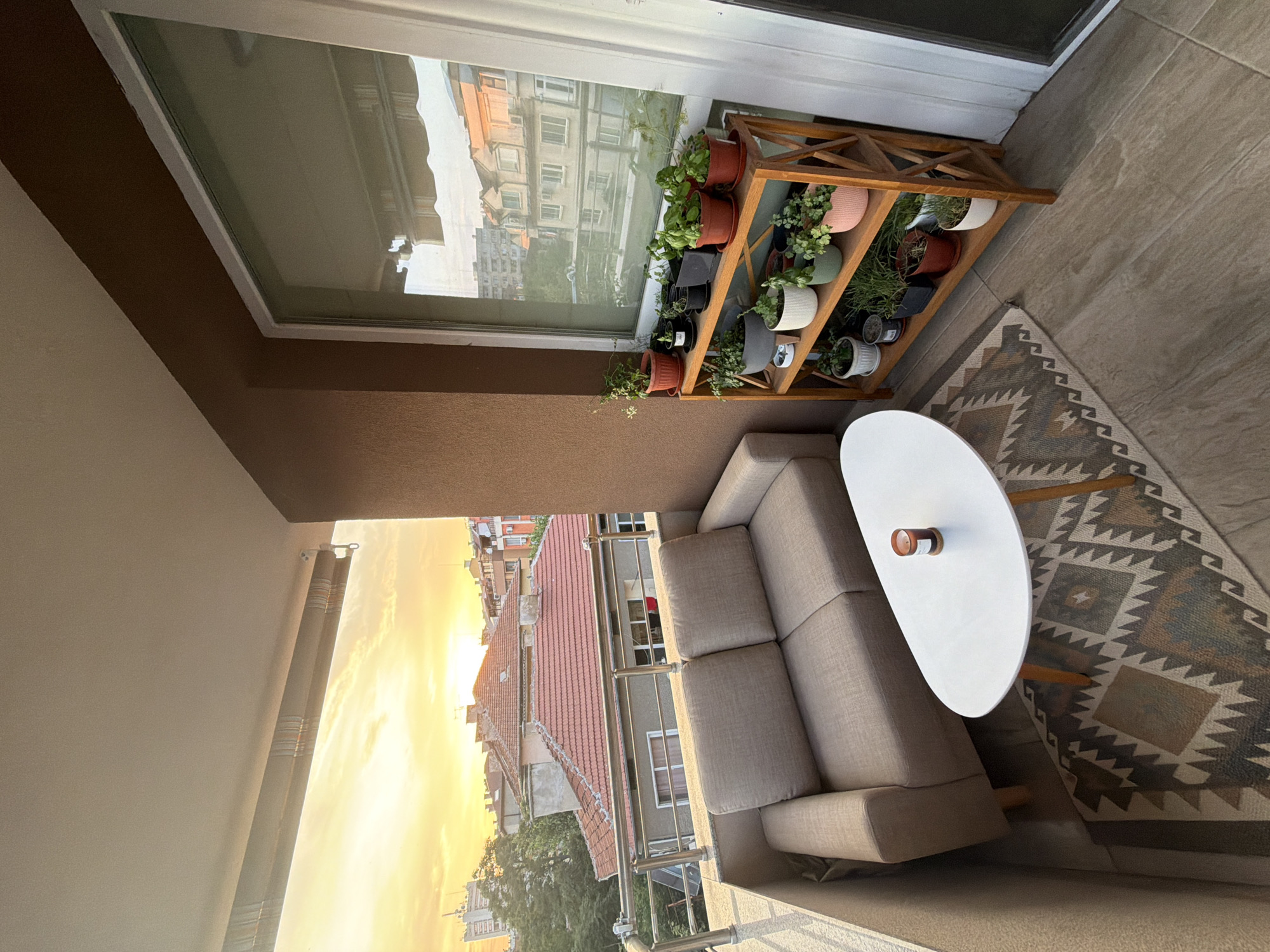 The image shows a balcony furnished with a two-seater sofa, a white oval coffee table with a candle on it, and a wooden tiered plant shelf filled with various potted plants. The balcony overlooks a city scene with buildings and rooftops bathed in the warm light of a sunset. A patterned rug is placed on the balcony floor.