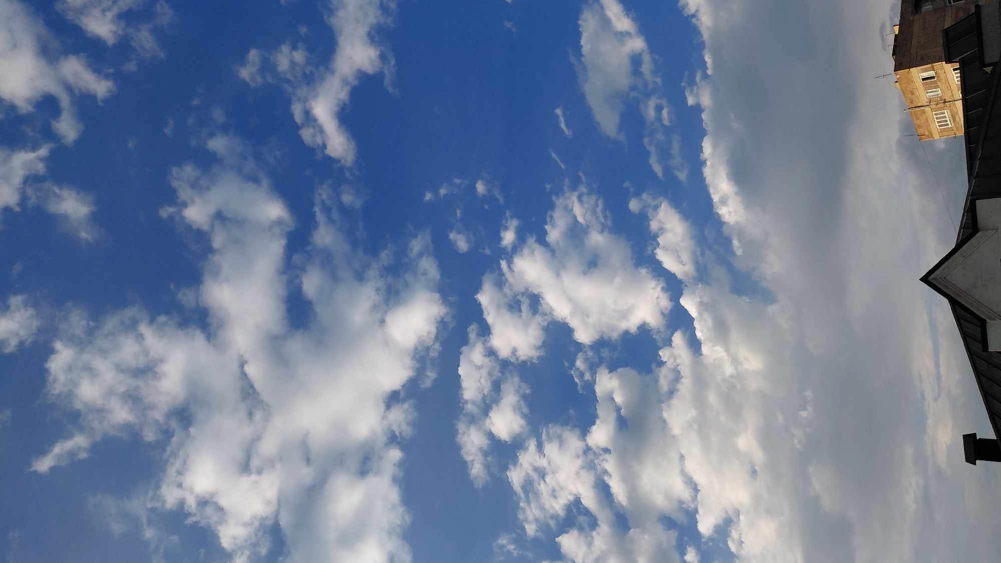 The image depicts a clear blue sky filled with scattered white cumulus clouds, suggesting fair weather. In the lower portion of the image, the roofs of two buildings are visible. The building on the left has a dark, sloped roof with a prominent chimney. To the right of this building, a multi-story brick building with windows can be seen. The scene appears to be an exterior view from a low vantage point, looking upwards towards the sky and capturing the tops of structures. The lighting suggests it might be late afternoon or early morning, given the angle of the light. The location is identified as Abovyan, Armenia.