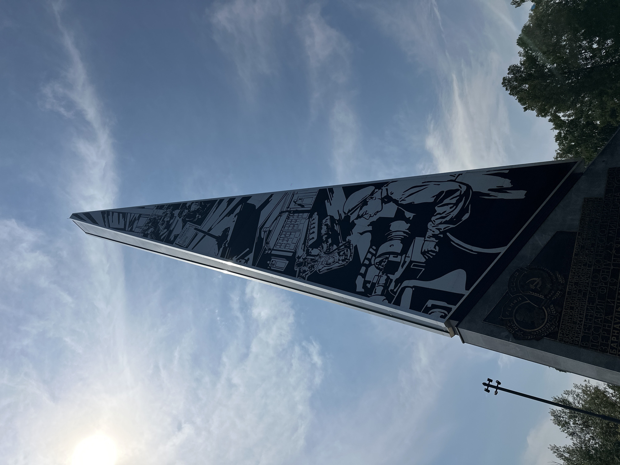 The image shows a view of the Monument to the Workers of the Altai Aviation Production Association named after Yu.A. Gagarin in Barnaul, Russia. The monument is a tall, obelisk-shaped structure with black and white engravings depicting scenes of industrial production and labor, as well as figures in profile. One prominent engraving shows a person operating what appears to be machinery. The base of the monument has a dark stone plaque with inscriptions in Russian and Soviet-era insignia, including the hammer and sickle. The monument is set against a bright blue sky with wispy clouds and the sun is visible in the upper left corner. Trees are visible on the right and left sides of the image, and a lamppost with multiple lights stands to the left of the monument. The perspective is from a low angle, looking up at the monument.