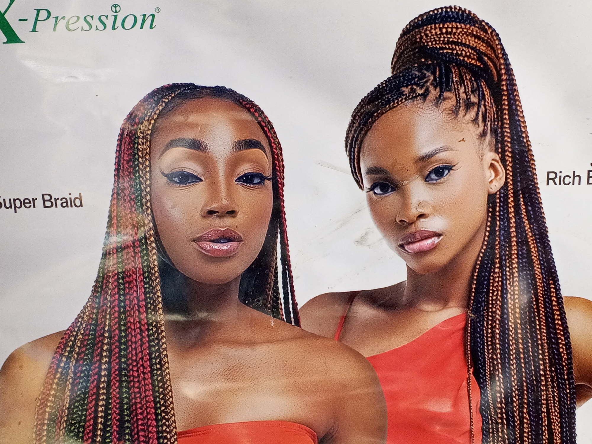 The image shows two women posing for a promotional advertisement for "X-Pression Super Braid" hair extensions.
The woman on the left has dark skin and shoulder-length box braids in varying shades of red, green, and black. She is wearing a red strapless top and heavy makeup, including eyeliner and lip gloss. She is facing forward.
The woman on the right has dark skin and long box braids, styled in a high ponytail with black and brown strands. She is wearing a red one-shoulder top. Her makeup is similar to the other woman's. She is facing forward with her head turned slightly to the left.
The background is white. Text is visible at the top left which reads "X-Pression". The other partial text in the image cannot be determined.
There is no explicit indication of the city of Gwong, Nigeria, or any recognizable environmental elements. The setting appears to be a studio or backdrop intended for product advertisement.