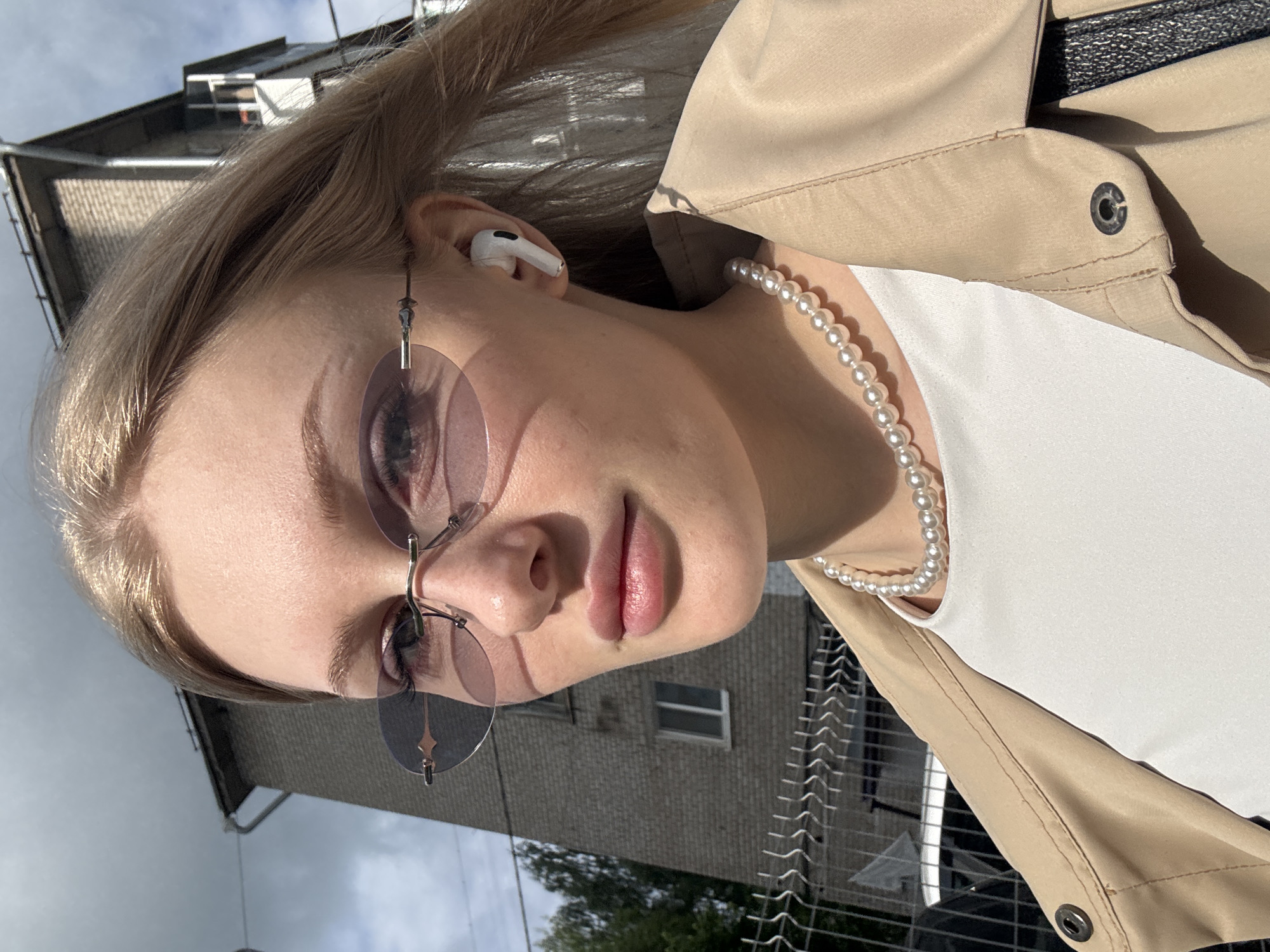 The image is a portrait-style shot of a young woman, taken from a slightly low angle, featuring her upper body and face. Her head is tilted, and the photo is rotated, showing her looking towards the viewer. She has light-colored hair, possibly blonde or light brown.
She is wearing rimless sunglasses with lightly tinted, possibly pink or purple, lenses. A white Apple AirPod is visible in her right ear. Around her neck, she wears a delicate pearl necklace. Her attire includes a light beige or tan trench coat or jacket, worn over a white top.
The background, largely out of focus, reveals an urban setting. A multi-story brick building, possibly residential or commercial, is visible behind her. A metal fence, likely chain-link or similar, runs along the bottom and side of the frame, suggesting a boundary or a public space like a sidewalk or park. Patches of green foliage or trees can be seen in the distance. The sky above is overcast or cloudy, indicating natural daylight without direct sunlight.
The woman appears to be posing for the camera, possibly taking a selfie, with a neutral to slightly serene expression. Her lips are softly parted. The time of day is clearly daytime. No visible text is present in the image. The provided location context for the scene is Perm, Russia.