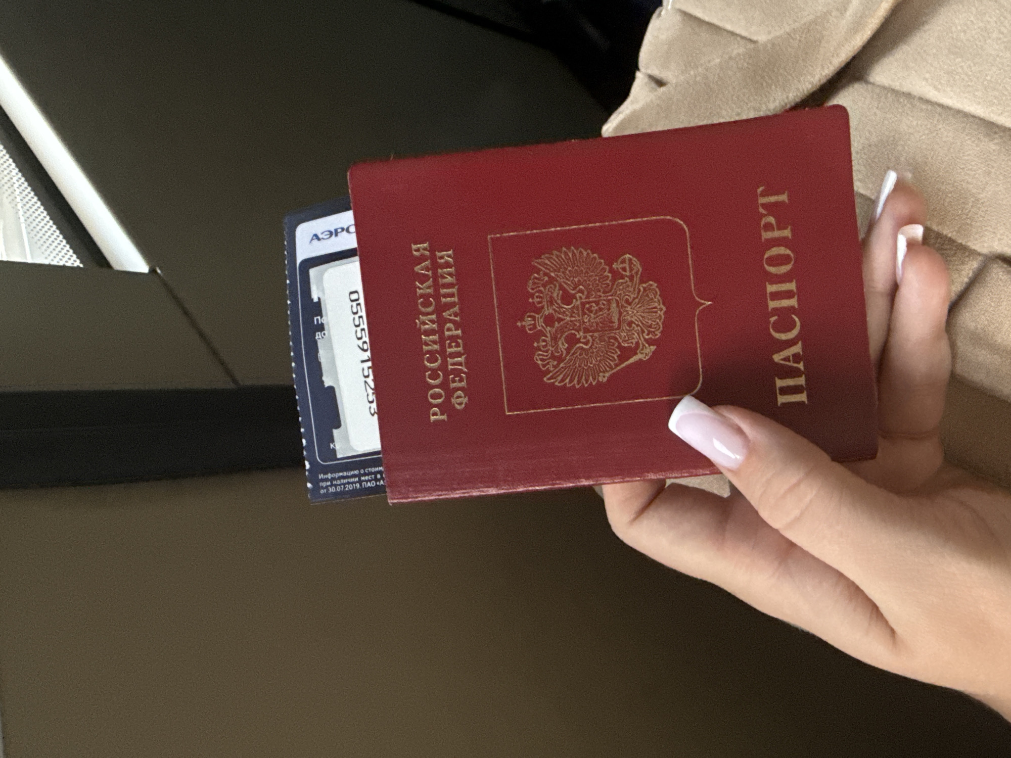 A woman's hand holds a Russian passport in its red cover, with the gold emblem of the Russian Federation visible on the front. A card with "АЭРО" and the number "0555915253" printed on it is partially inserted into the passport. The Russian words "РОССИЙСКАЯ ФЕДЕРАЦИЯ" and "ПАСПОРТ" are displayed on the cover. The text "Информацию о стоим при наличии мест в с от 30.07.2019. ПАО «Α По до" is on the card. The woman has manicured nails with white tips. The background includes a dark surface and a beige garment. The location is in Russia, as suggested by the passport. The card is from the Moscow airport, which is near the city of Khimki.