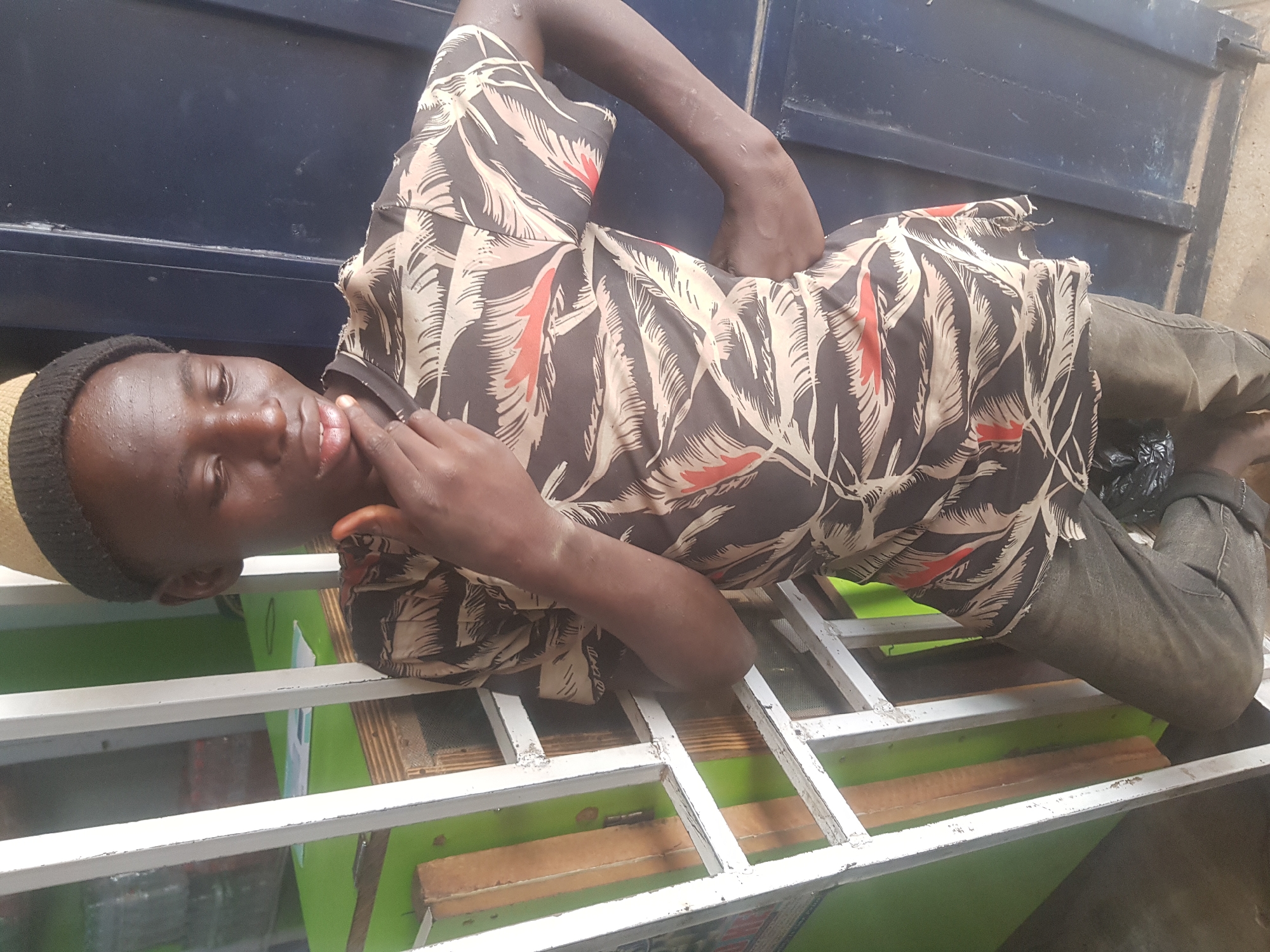 The image depicts a young man lying on a white, metal grid. He is wearing a black and cream-colored patterned shirt, dark pants, and a small tan hat. He has his right hand near his mouth and is looking towards the camera. Behind the grid is a green and brown surface. In the background, there is a dark blue metal panel. The location is identified as Kufena, Nigeria. There are no visible landmarks to pinpoint the specific location.