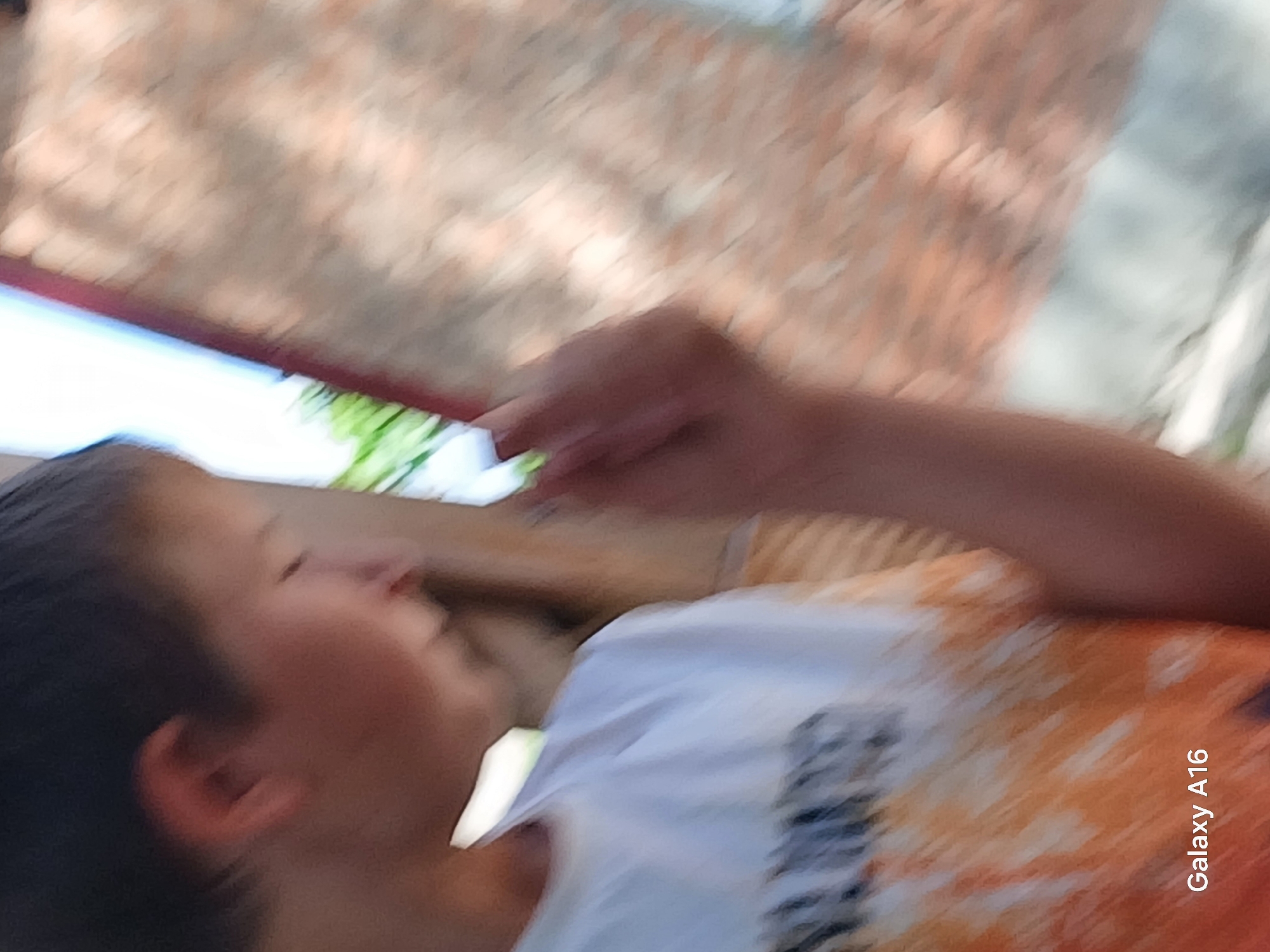 This is a poorly focused, medium shot photograph taken outdoors. A young person is visible from the side, facing to the left. They are wearing a white t-shirt with an orange and black abstract design. The person's left ear and the side of their face are visible. The background consists of a brick wall and some greenery, suggesting an outdoor setting. The bottom left corner of the image displays "Galaxy A16," indicating the device used for capture. The image lacks sufficient detail for a precise forensic analysis or determination of location within Ustyvytsia, Ukraine.
