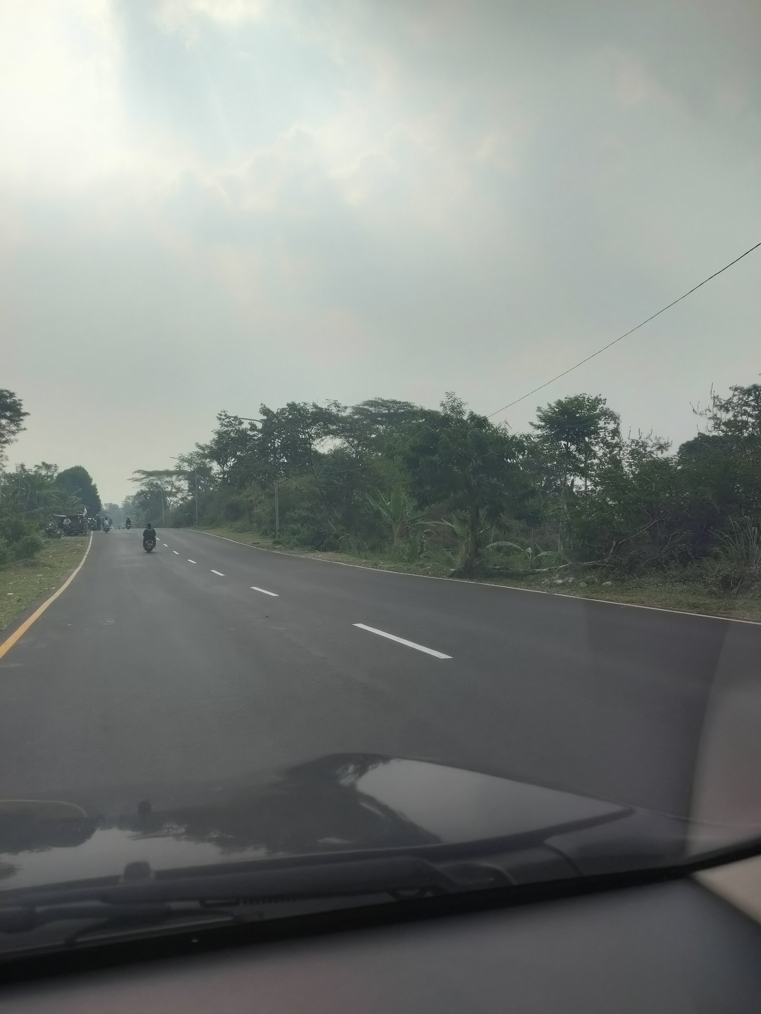The image shows a view from a vehicle driving on a two-lane asphalt road. The road has white dashed lines indicating lanes. On the left side of the road, there is a yellow line marking the shoulder, followed by a grassy area and then trees. On the right side of the road, there is a shoulder with grass and dense vegetation, including trees and banana plants. The sky is overcast with diffused sunlight. Several motorcycles are visible on the road. One motorcycle is in the foreground, ridden by a person. In the distance, on the left side of the road, a group of people and motorcycles are gathered. The overall impression is a rural or semi-rural setting on a highway, potentially in the city of Subang, Indonesia, given the vegetation and prevalent use of motorcycles. The front of the vehicle, including the dashboard and windshield wipers, are visible in the bottom portion of the image.
