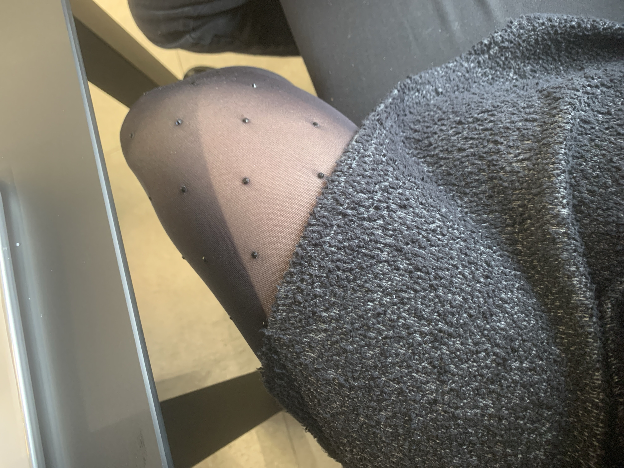 A close-up shot from above shows a person's leg covered in black patterned tights and part of a dark grey, textured sweater or blanket. The leg is bent, suggesting the person is sitting. A portion of a grey, possibly metal or plastic, object is visible on the left edge of the frame. The background is out of focus and shows a light-colored flooring and a hint of what appears to be furniture legs. No identifying features suggest the location is specifically Izhevsk, Russia.