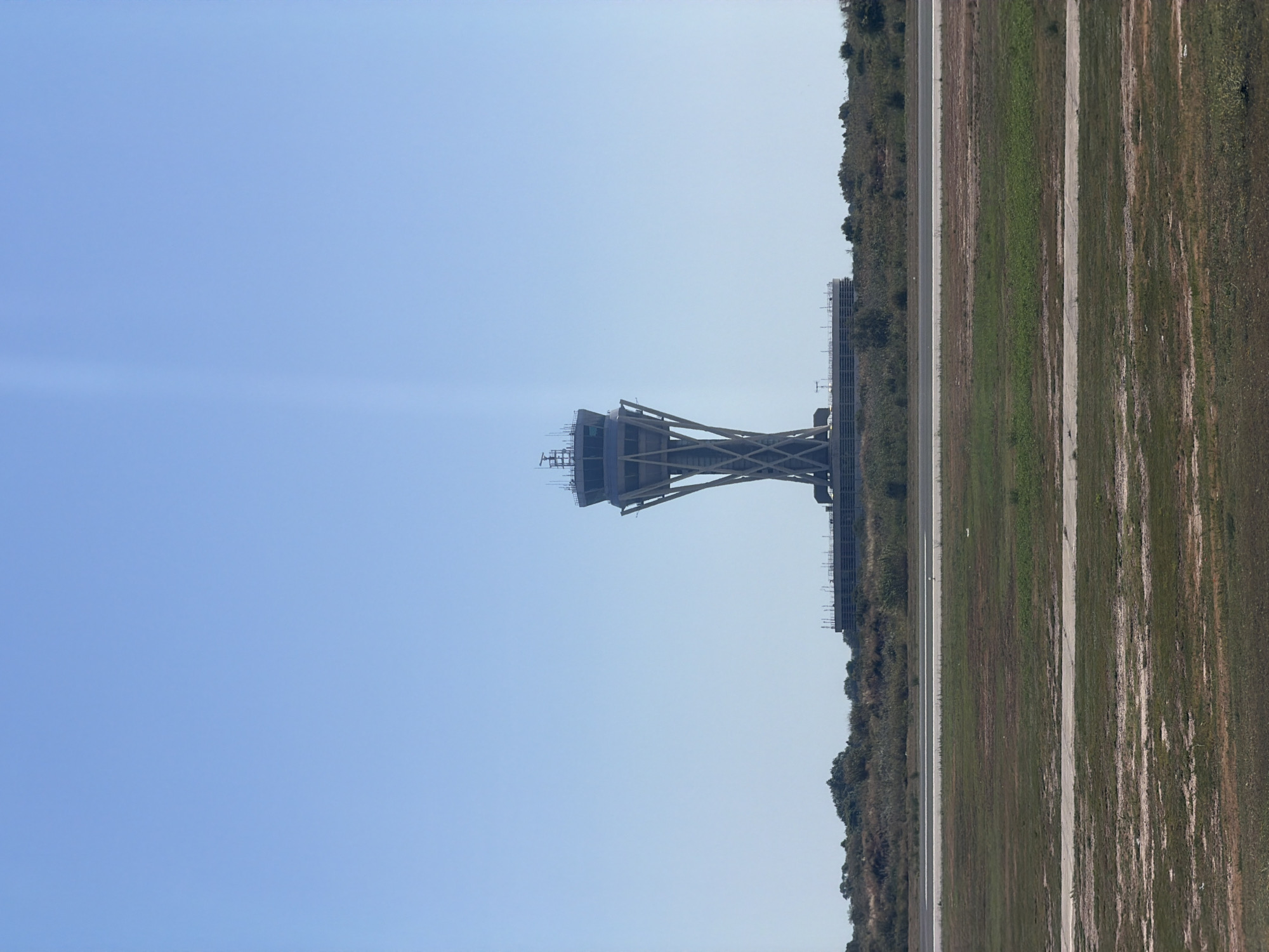 An air traffic control tower stands prominently against a clear blue sky. The tower, located in El Prat de Llobregat, Spain, features a distinctive X-shaped base supporting a cylindrical control cabin at the top. The surrounding environment consists of a grassy field in the foreground, with a paved runway or road visible beyond it. Further back, there is a line of trees and bushes, and behind that, a low-lying building structure. The overall scene depicts an airport environment under a bright, cloudless day.