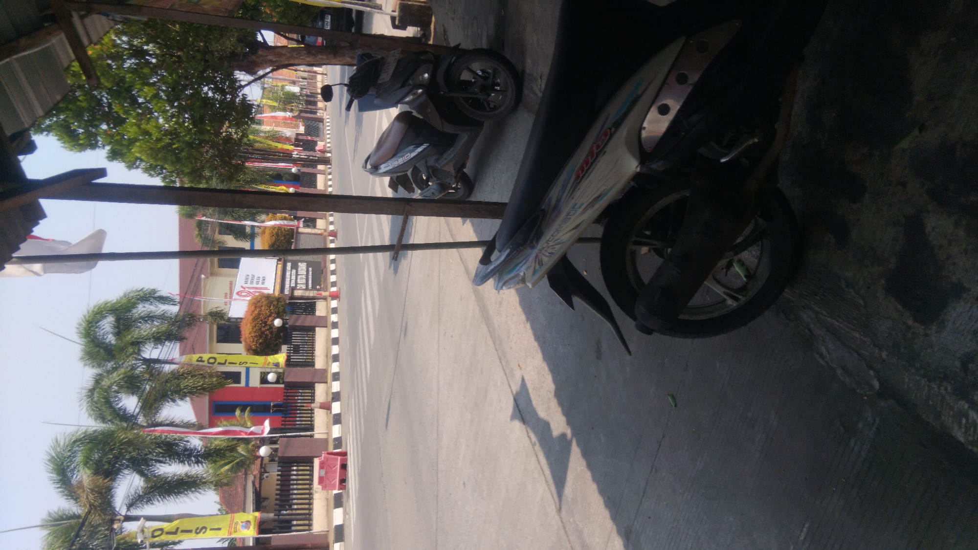 The image depicts a street scene in Proyonangg, Indonesia. In the foreground, two motorcycles are parked on a paved surface, one in front of the other, partially obscuring the view of the street. The motorcycle in the foreground is white and blue with the word "AUTOMATIC" visible on its side. The second motorcycle, parked slightly behind it, is dark gray.
Beyond the motorcycles, a street with pedestrian crossing marks stretches across the frame. On the far side of the street, a building with red and yellow accents is visible, possibly a government or public institution, as indicated by flags and banners that read "POLISI" (police). Palm trees and bushes are planted in front of the building. The sky is clear and bright, suggesting a sunny day. The overall environment appears to be an urban or suburban setting.