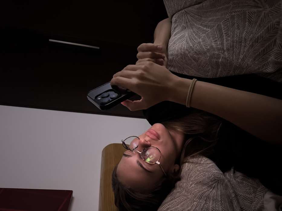 A woman with glasses lies down, holding a smartphone above her. She rests on a patterned pillow, with a dark ceiling and a light fixture visible above. Her arm with a bracelet extends towards the phone. The background includes a white wall and a wooden headboard. The scene is dimly lit, creating a cozy and intimate atmosphere.