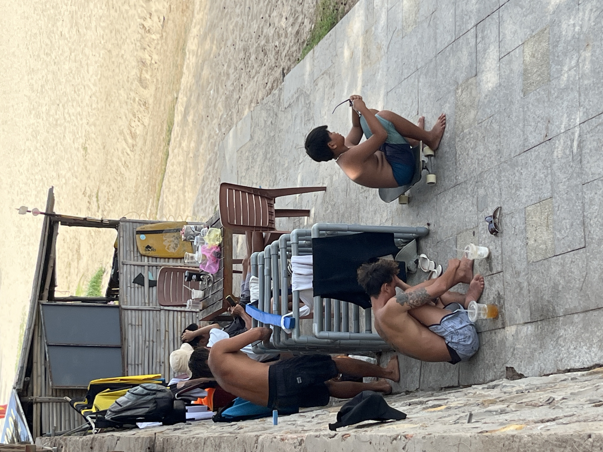 In Phu Thuy Ward, Vietnam, on a paved area adjacent to a sandy beach, a group of individuals are gathered near a bamboo structure. In the foreground, a young male with dark hair and wearing multi-colored board shorts sits on a skateboard, holding sunglasses in his right hand and looking down. To his left, another young male, shirtless and with tattoos on his arm and torso, is crouched down, with his head bowed and right hand covering his face. In front of him are two disposable cups, one with a straw and a light-colored liquid, and a pair of flip-flops. To the left of these two individuals, a shirtless male wearing dark shorts stands facing away from the camera, his back to the viewer, with his right hand touching his head. A rack with various clothing items hangs behind him. Further in the background, a wooden table with chairs is set up outside the bamboo structure. Several cups and pink and yellow plastic bags are on the table. Beyond the paved area is a sandy beach with sparse vegetation.