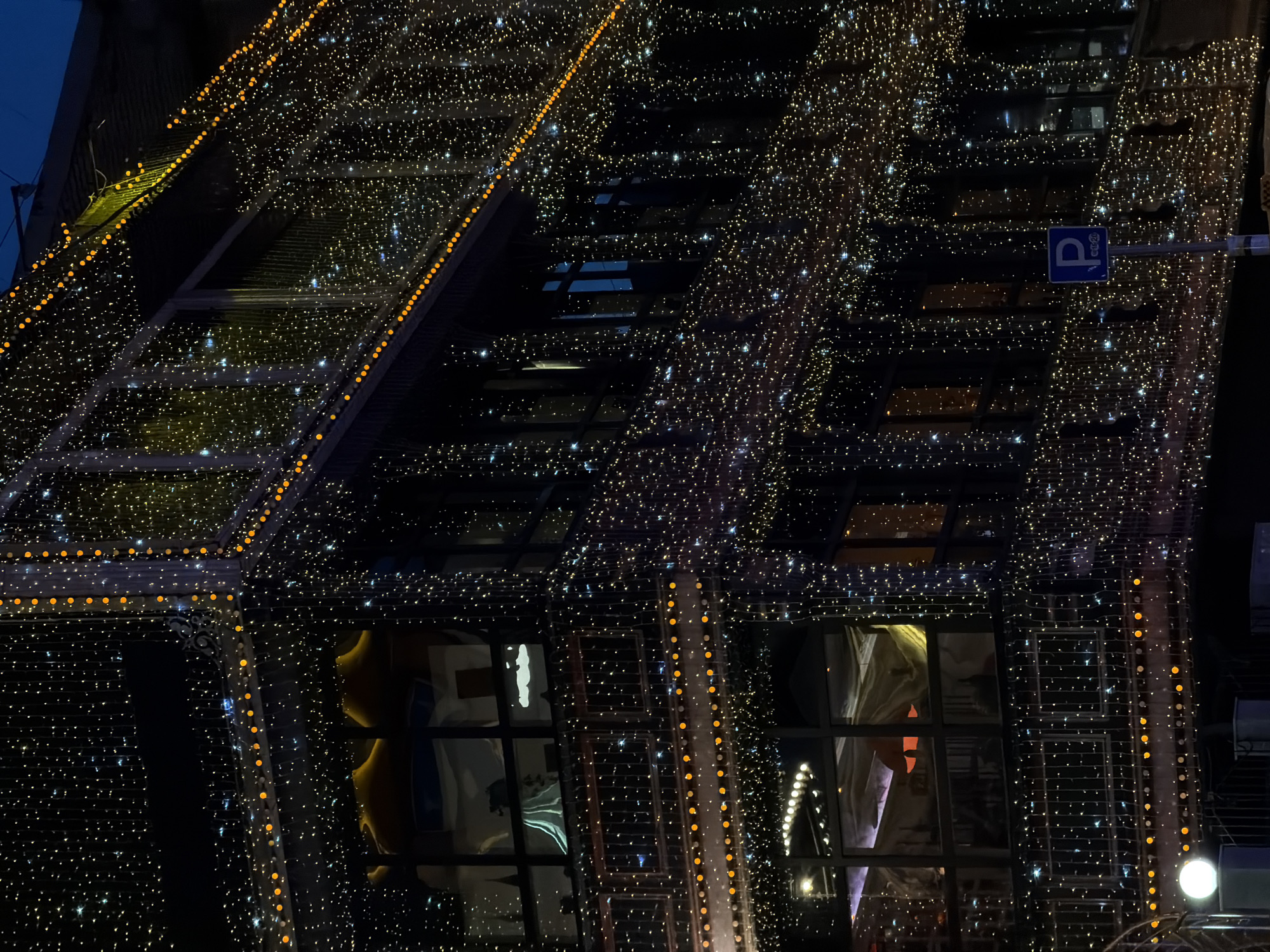 The image depicts a multi-story building in Moscow, Russia, at night, extensively decorated with festive lighting. The building's dark facade is predominantly covered by dense, vertical strings of small, warm-toned string lights, creating a curtain-like effect across its front and right side. Larger, orange-yellow circular lights accentuate architectural features, outlining window frames, the perimeter of the building, and railings on the balconies.
Through the large, dark-framed windows on the upper-left floor, illuminated interior elements, possibly curved seating or decor, are visible. On the middle-left floor, interior lighting reveals faint reflections and obscured shapes, including a white, cloud-like decoration and a vertical white screen or curtain within the windows. Windows on the right side reflect the outdoor lights and display dark interiors.
In the lower-right foreground, a blue rectangular road sign with a white 'P' symbol for parking and the text "еu 24" (Russian shorthand for "daily 24 hours") is mounted on a metal pole. In the lower-left corner, a spherical white lamp sits atop a dark pillar, emitting a soft glow. The sky above is a dark blue, indicative of dusk or nighttime. No individuals or specific actions are discernible in the scene.