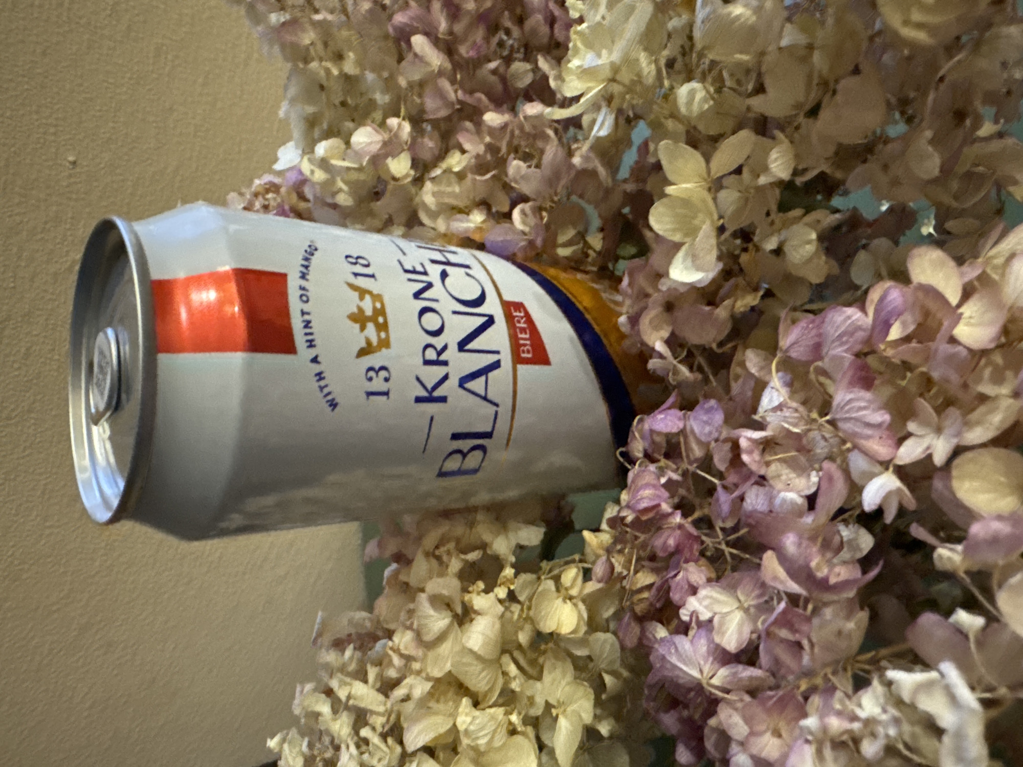 A can of Kronenbourg Blanc 13/18 beer is partially obscured by dried hydrangea flowers of varying shades of cream, beige, and light purple. The beer can is silver with a red stripe near the top, and a gold and blue section towards the middle. The scene appears to be indoors. The location of the image cannot be determined to be Votkinsk, Russia.