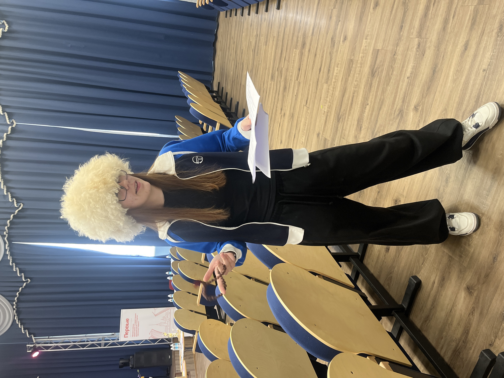 A young person stands in an auditorium or conference hall, likely in Novoe Devyatkino, Russia. The individual, appearing youthful with long brown hair and glasses, is wearing a distinctive white, fluffy hat, a blue and white track jacket over a black top, black wide-leg pants, and white sneakers with blue detailing.
In their right hand, they hold a folded piece of white paper. Their left hand holds a string of dark beads, possibly prayer beads (tasbih), extended over the back of a nearby chair. The person has a slight, pleasant smile and looks towards the left of the frame.
The setting consists of an empty hall with rows of chairs featuring light wooden backs and blue upholstered seats, arranged on a wooden floor. Some chairs have unique golden, wing-like decorative elements on their backs. Blue curtains line the back wall. To the far left, a stage area is visible with sound equipment, including a speaker and a metal truss structure with lights. A vertical banner on the stage displays the Russian word "Первые" (Pervye), meaning "The First ones" or "The Pioneers," followed by smaller text indicating "общественная организация" (public organization) and other details that are partly obscured. Small bottles of water are visible on some of the chairs. The room is well-lit, suggesting an indoor event venue.
