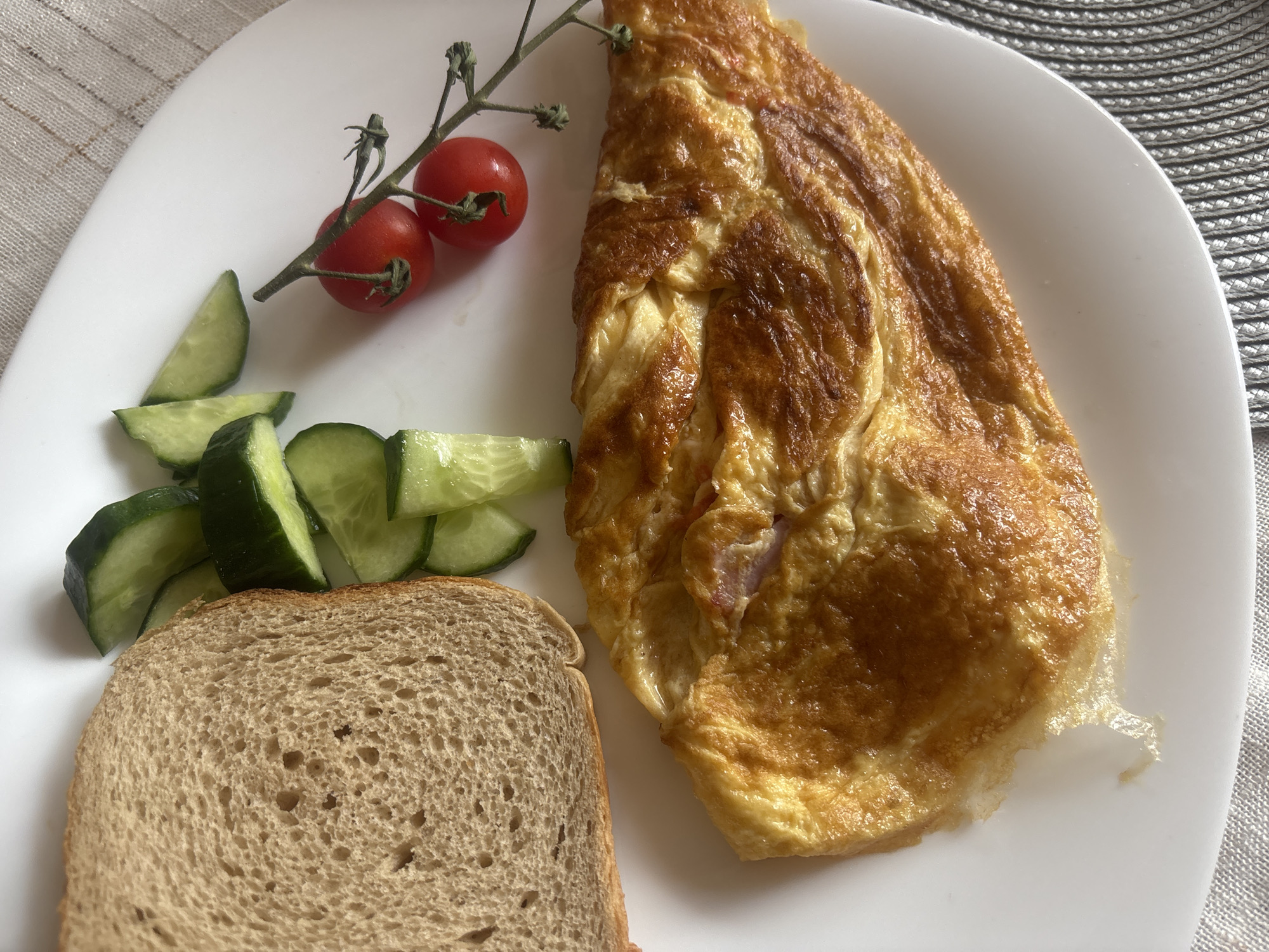 An overhead shot shows a white plate with a breakfast. On the plate are a slice of whole wheat toast, sliced cucumber, cherry tomatoes on the vine, and an omelet. The food is positioned on a woven placemat, which rests on a table. The image lacks any contextual clues that would allow for identification of a specific location within Plovdiv, Bulgaria, or any other city.