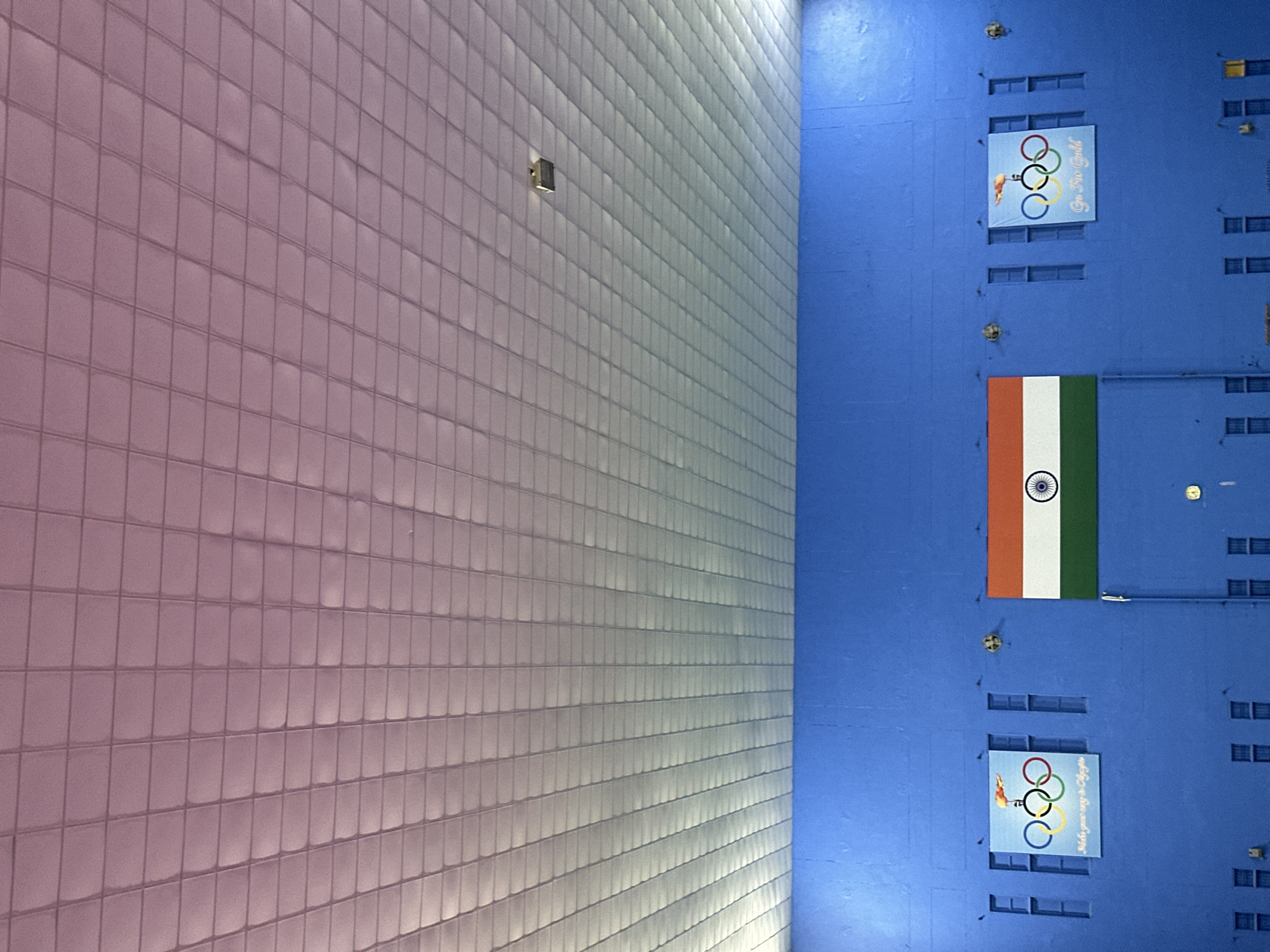 The image shows a wide shot of the interior of what appears to be an indoor sports facility. The dominant feature is a bright blue wall that takes up the lower half of the image. On this wall, there are several decorative elements and architectural features. Centered on the wall is the Indian flag, with its tricolor bands of saffron, white, and green, and the Ashoka Chakra in the center of the white band. To the left and right of the Indian flag are two banners featuring the Olympic rings, with flames above them. The banner on the left has text that reads "Make your way to Olympics," and the banner on the right has text that reads "Go For Gold." There are also multiple vertical, rectangular indentations or windows spaced along the blue wall.
The upper half of the image is dominated by a white, tiled ceiling that recedes into the distance. The tiles are arranged in a grid pattern, and there is a subtle gradient of light and shadow across the ceiling, with a hint of pinkish light at the top, possibly from lighting fixtures. A single, square light fixture is visible on the ceiling in the upper right quadrant of the image. The overall impression is one of an indoor space designed for sports or athletic training, decorated with patriotic and motivational imagery related to the Olympics. The image does not contain any people. The location is identified as Unknown, Unknown.