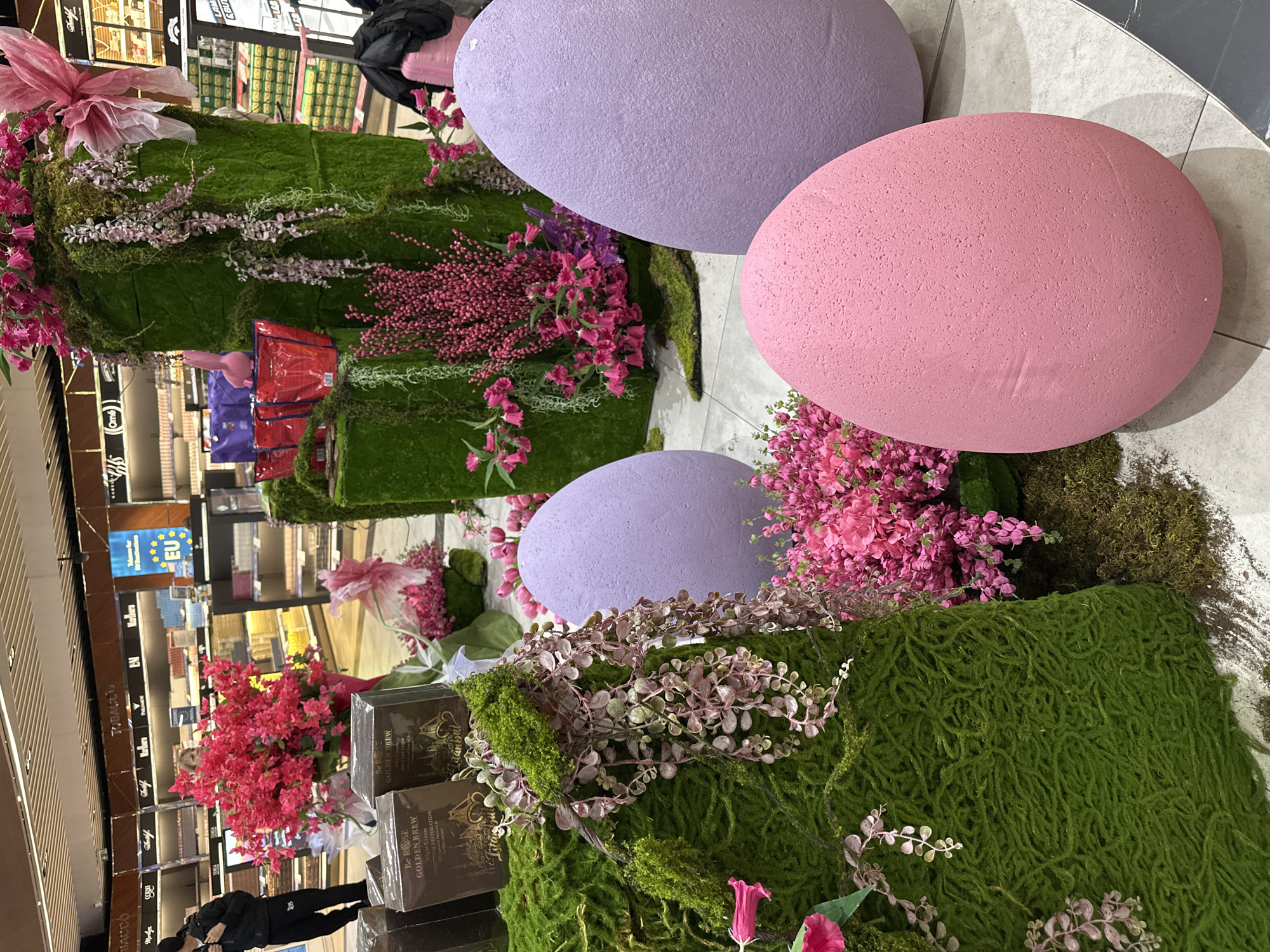 Here's a detailed description of the image:
**Scene:** The image appears to be taken inside a duty-free store or similar retail environment, possibly at an airport. The primary focus is on an elaborate Easter display, which incorporates oversized, pastel-colored (pink and lavender) eggs, lush green moss, and various pink and purple floral arrangements.
**Objects:**
* **Easter Eggs:** Two large lavender eggs and one large pink egg dominate the display.
* **Greenery:** The eggs and other objects are surrounded by vibrant green moss, creating a natural, garden-like setting.
* **Flowers:** Various pink and purple flowers are integrated into the display, adding color and texture.
* **Retail Products:** Partially visible are boxes of "Be ROSE GOLDEN BREW" night cream, suggesting the display is within a store selling beauty or personal care products.
* **Shelves and Products:** In the background, there are shelves filled with various products, including tobacco products (brands like "Marilyn", "Dolf"), and other items suggesting a duty-free environment.
* **Signage:** A sign labeled "EU" is visible on one of the shelves.
**People:**
* A person is partially visible towards the lower-left corner of the frame. They appear to be wearing dark clothing.
**Environment:** The store is brightly lit and appears to be well-organized. The flooring is light-colored, possibly tile or polished concrete.
**Location Clues:**
* The product "Be ROSE GOLDEN BREW" offers the primary indicator.
* However, I don't have specific information to confirm if this particular brand is exclusive to Bulgaria or Sofia. Further research on the product's distribution or store chains in the background might help pinpoint the location more accurately.