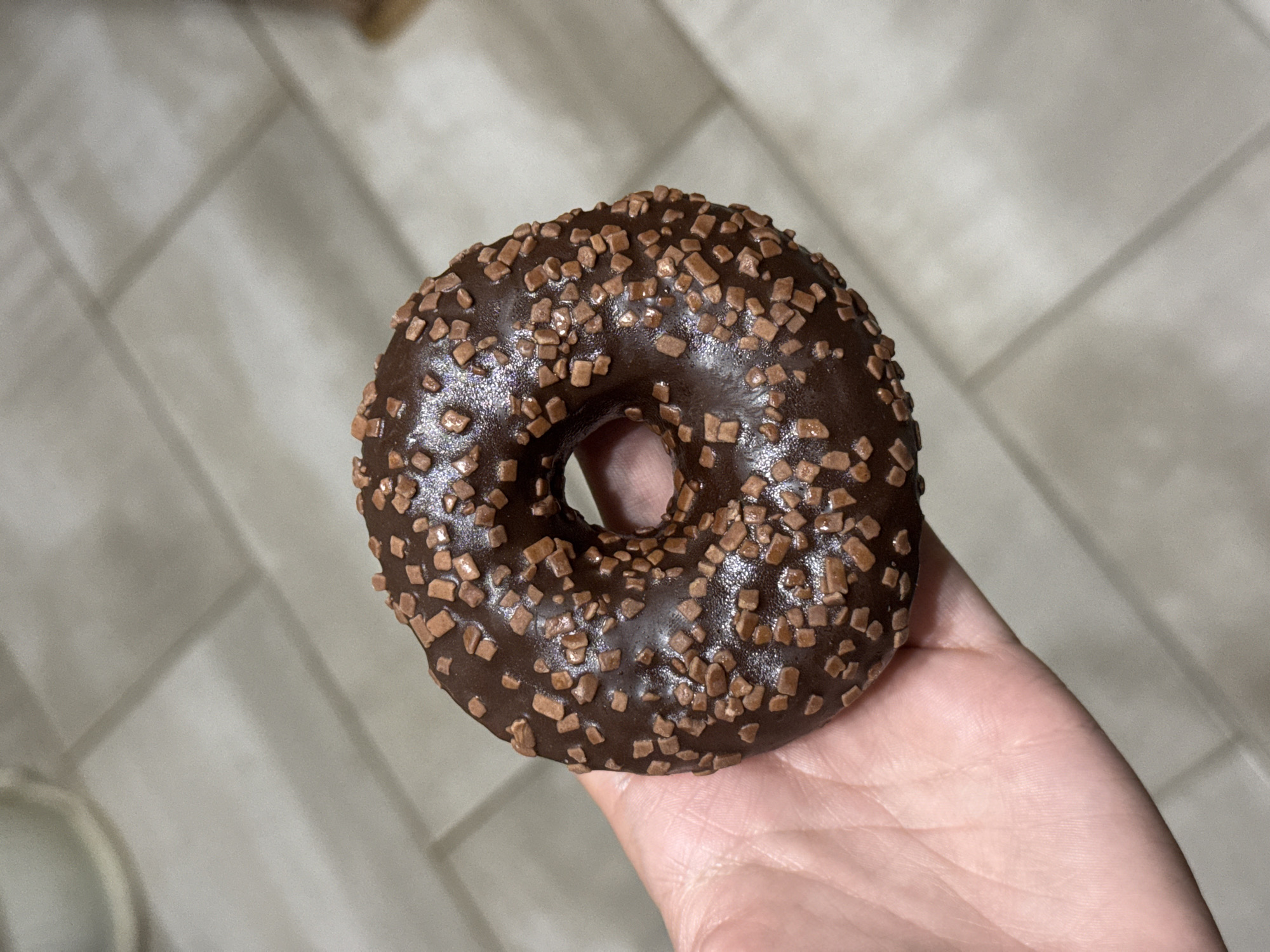 The scene is located in Stara Zagora, Bulgaria. A single, round doughnut is centered in the frame, held by an unidentified individual's left hand. The doughnut features a dark brown, glossy chocolate glaze uniformly covering its surface, generously topped with light brown, irregularly shaped, granular sprinkles. The doughnut has a distinct circular hole in its center. The hand, visible from the palm and fingers, supports the doughnut from underneath. The background consists of light grey or beige rectangular tiles, arranged in a horizontal and vertical grid pattern, suggesting a floor or countertop surface. Shadows and reflections indicate an indoor setting with overhead lighting.