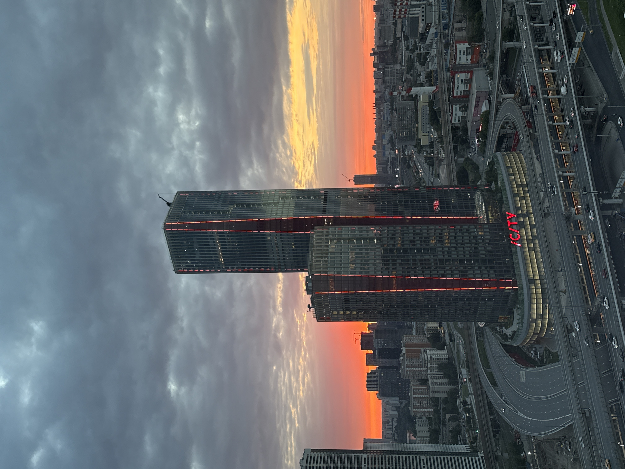 The image shows a panoramic view of the city of Khimki, Russia, during sunset. The sky is filled with dramatic clouds, with hues of orange and yellow from the setting sun peeking through. In the foreground, two prominent skyscrapers with illuminated red vertical lines stand tall. The larger skyscraper, on the right, is partially obscured by the clouds but its upper section is visible. The smaller, wider skyscraper to its left is more fully visible, and it features a circular base with illuminated signage. A complex network of elevated highways and roads surrounds the base of these buildings, with several cars visible on the roads, suggesting active traffic. Beyond the skyscrapers, a dense urban landscape of various buildings stretches into the distance, with more industrial structures and chimneys visible. The overall impression is that of a modern, bustling city at dusk.