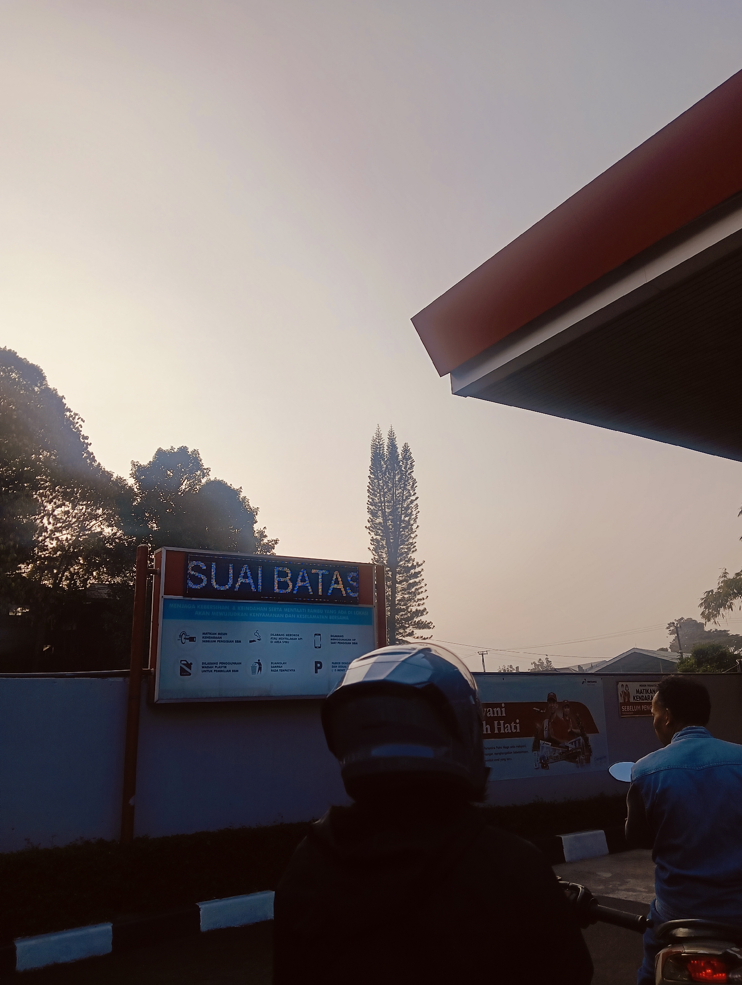 The image depicts a scene at a gas station, likely in Cisarua, Indonesia, during what appears to be early morning or late afternoon, given the soft, diffused light and hazy sky. In the foreground, two individuals on motorcycles are visible. The person on the left is seen from behind, wearing a dark helmet and jacket. The person on the right, also on a motorcycle and viewed from behind, is wearing a light blue denim shirt.
Dominating the mid-ground is a digital billboard displaying the text "SUAI BATAS" in large, illuminated letters, with smaller text below detailing regulations for maintaining cleanliness and order at the location. The billboard is mounted on a dark gray wall. To the left of the billboard, silhouetted trees with dense foliage are visible against the bright sky. A tall, slender pine tree stands to the right of the billboard.
In the background, several low-rise buildings are visible, along with more trees and utility poles. A portion of the gas station's red and white awning extends from the upper right of the frame. The ground is paved, with white lines indicating parking spaces. The overall atmosphere is calm and subdued.